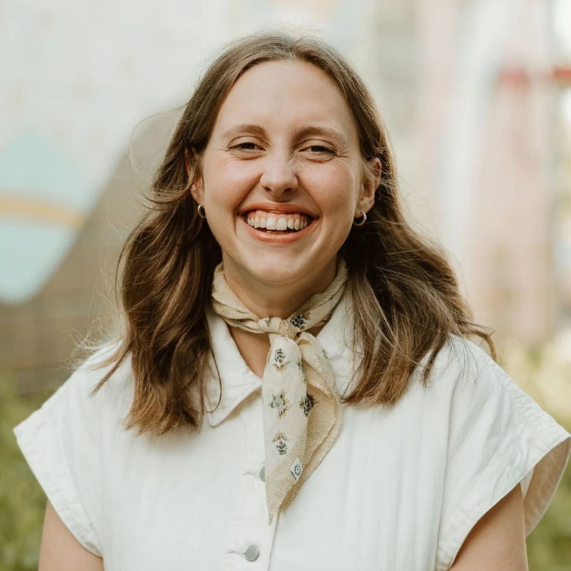 A headshot of a young woman with shoulder-length brown hair, smiling broadly at the camera. She wears a cream-colored shirt and a patterned neckerchief.
