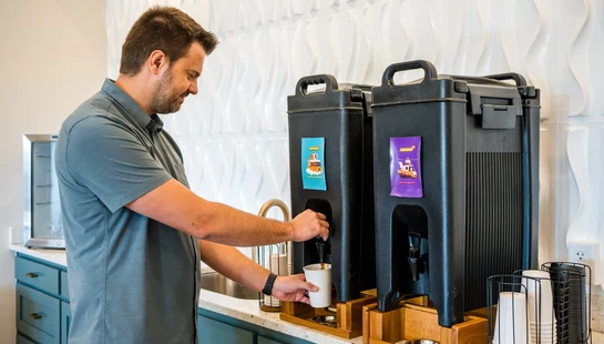 A man in a gray shirt pours coffee from a large black dispenser into a white mug at a modern beverage station with a textured white wall.