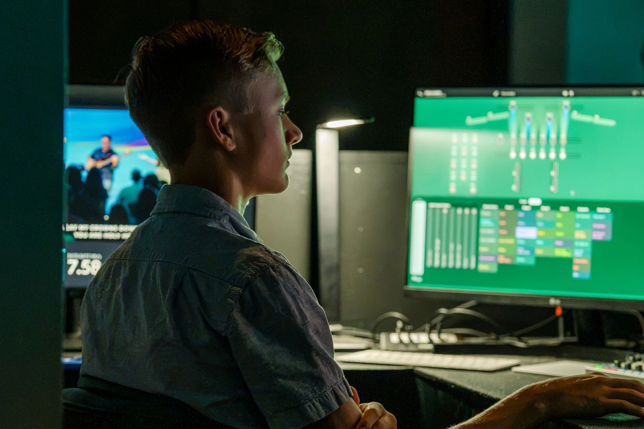 A young man sits in profile in a dimly lit room, focused on a large computer monitor displaying an audio or video editing interface for a live event.