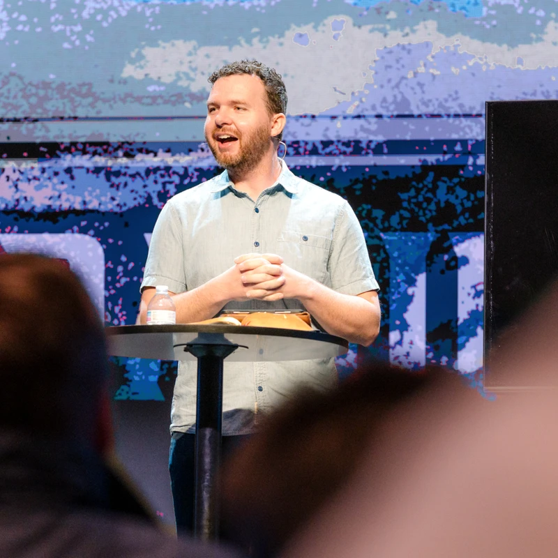 A bearded man in a light blue shirt stands at a lectern on stage, speaking to an audience. Behind him is a large screen with an abstract blue graphic.
