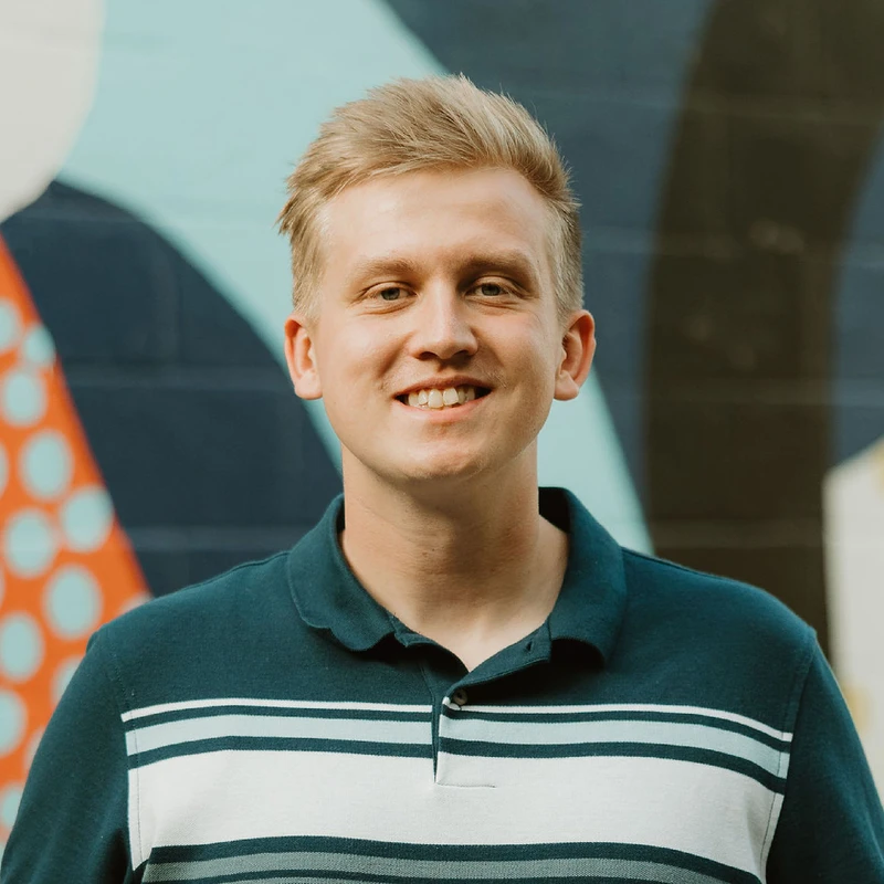 A headshot of a smiling young man with blonde hair wearing a striped polo shirt. He is standing in front of a colorful, abstract mural on a wall.