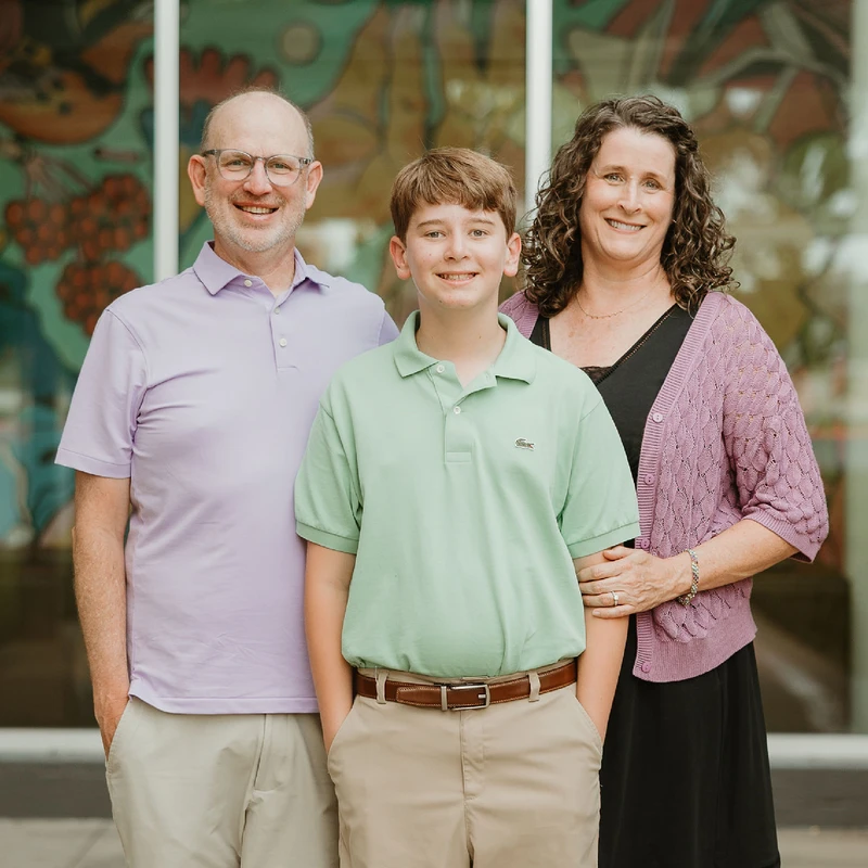A man, a woman, and their teenage son smile for the camera while standing together outdoors in front of a colorful building.