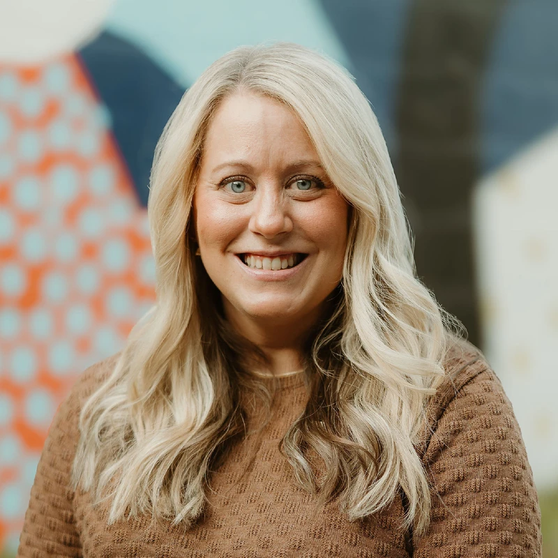 A headshot of a smiling woman with long blonde hair and blue eyes. She is wearing a brown sweater and standing in front of a colorful, abstract background.