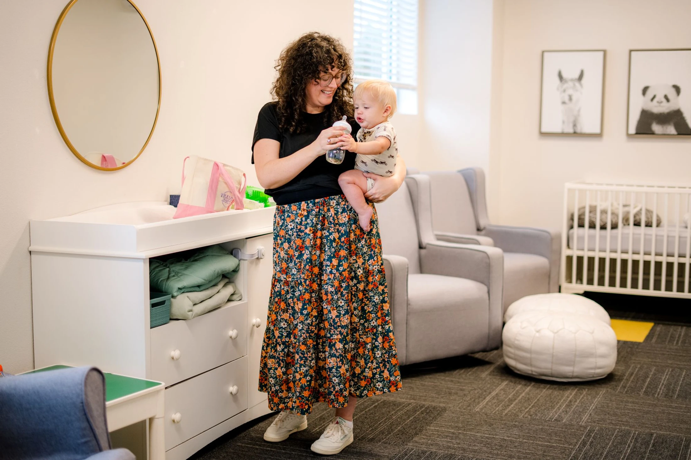 A smiling woman with curly hair and glasses holds a small baby in a nursery, offering them a bottle next to a white changing table.