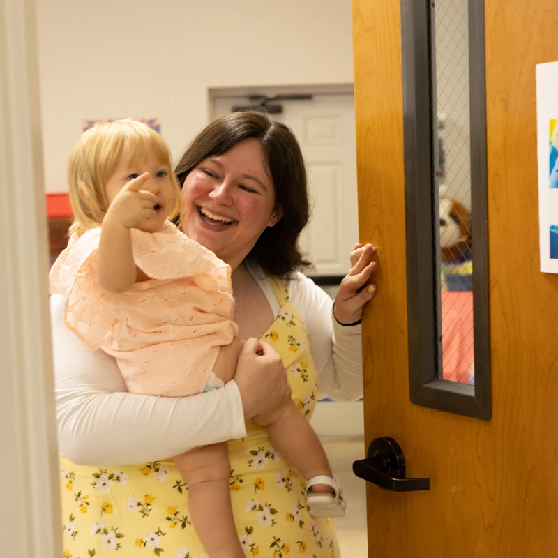 A happy woman with dark hair holds a small blonde toddler in her arms. The child points forward as the woman laughs, standing next to a wooden door.