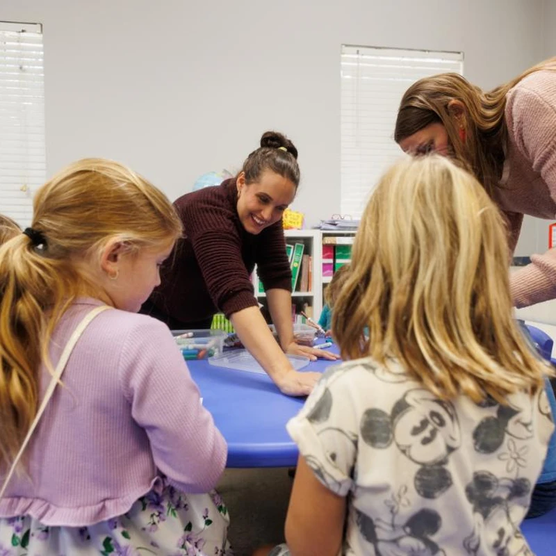 Two female teachers lean over a blue table, smiling and helping a group of young girls with an art project in a classroom.