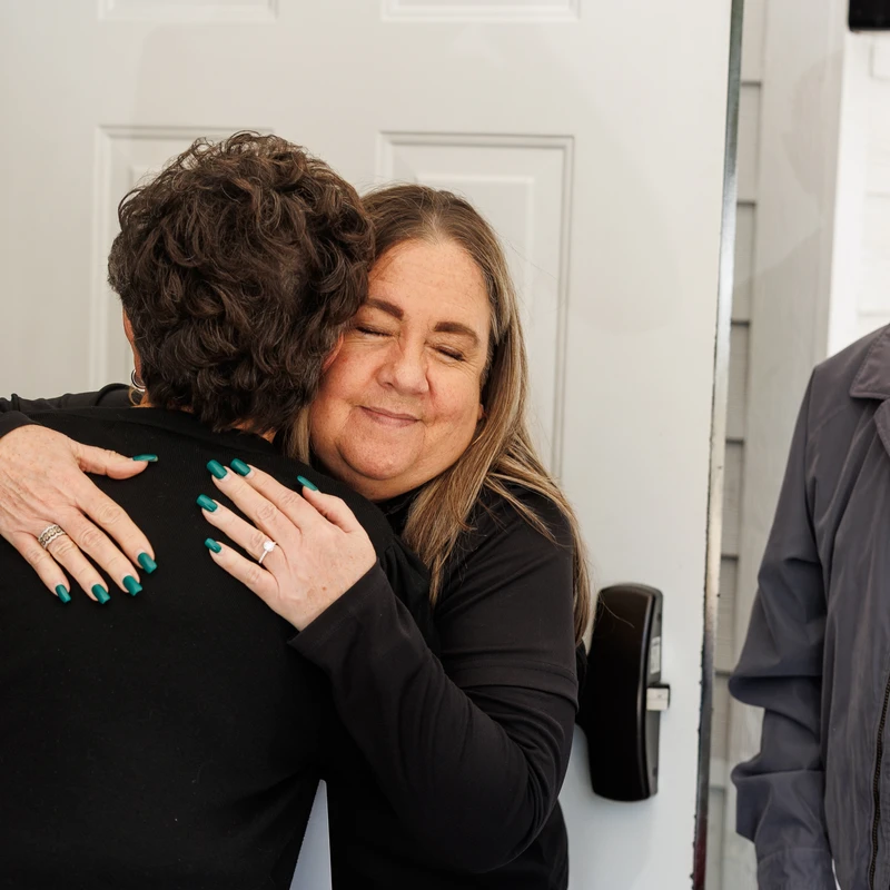 A middle-aged woman with her eyes closed smiles warmly as she hugs another person. An elderly man stands beside them, watching.