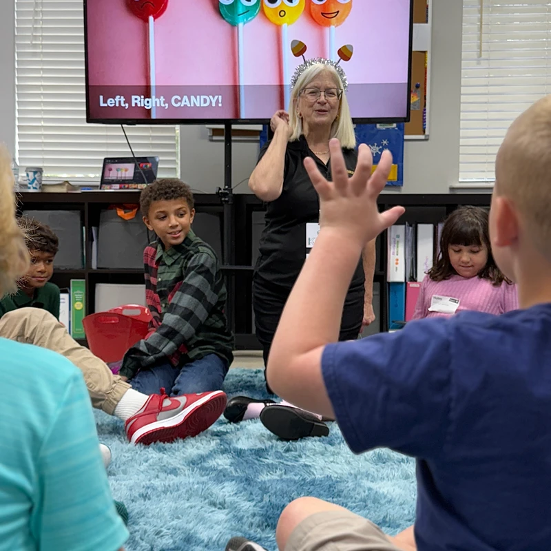 A teacher wearing a candy corn headband leads a lesson for children sitting on a blue rug, with a screen showing cartoon lollipops behind her.