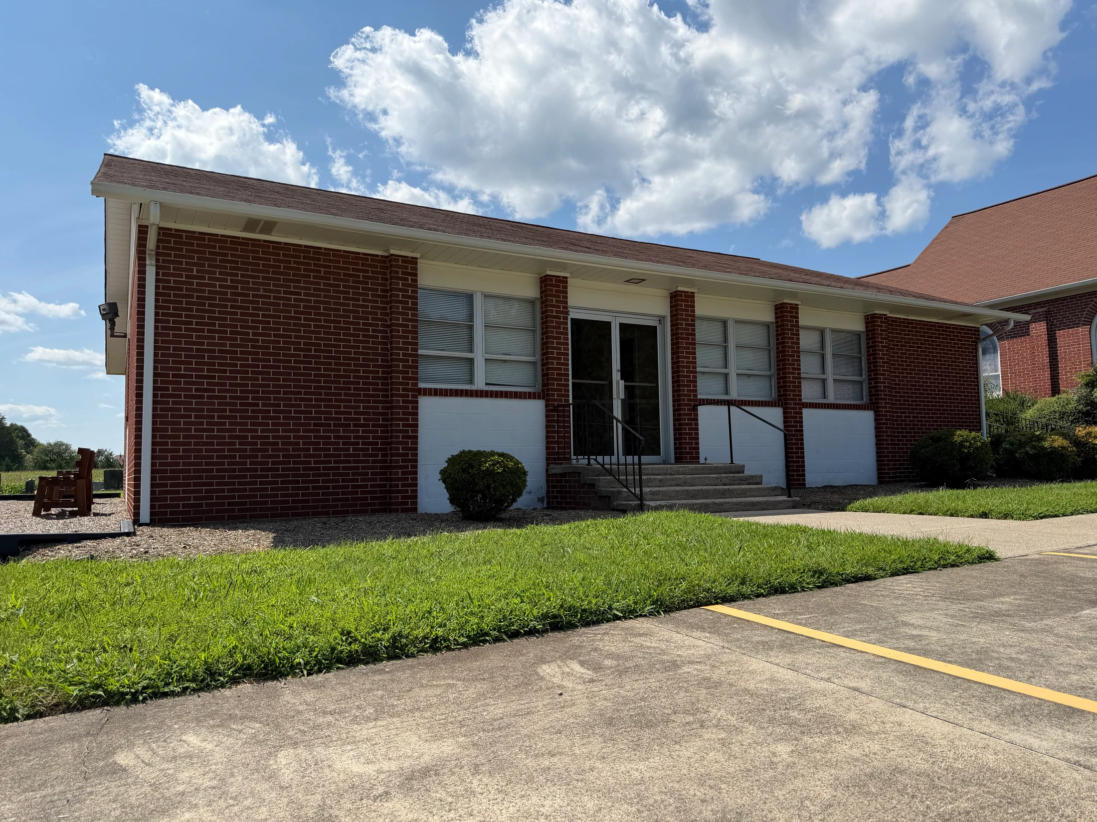 A one-story red brick building with a covered entrance and concrete steps, set against a bright blue sky with white clouds. A green lawn is in the foreground.