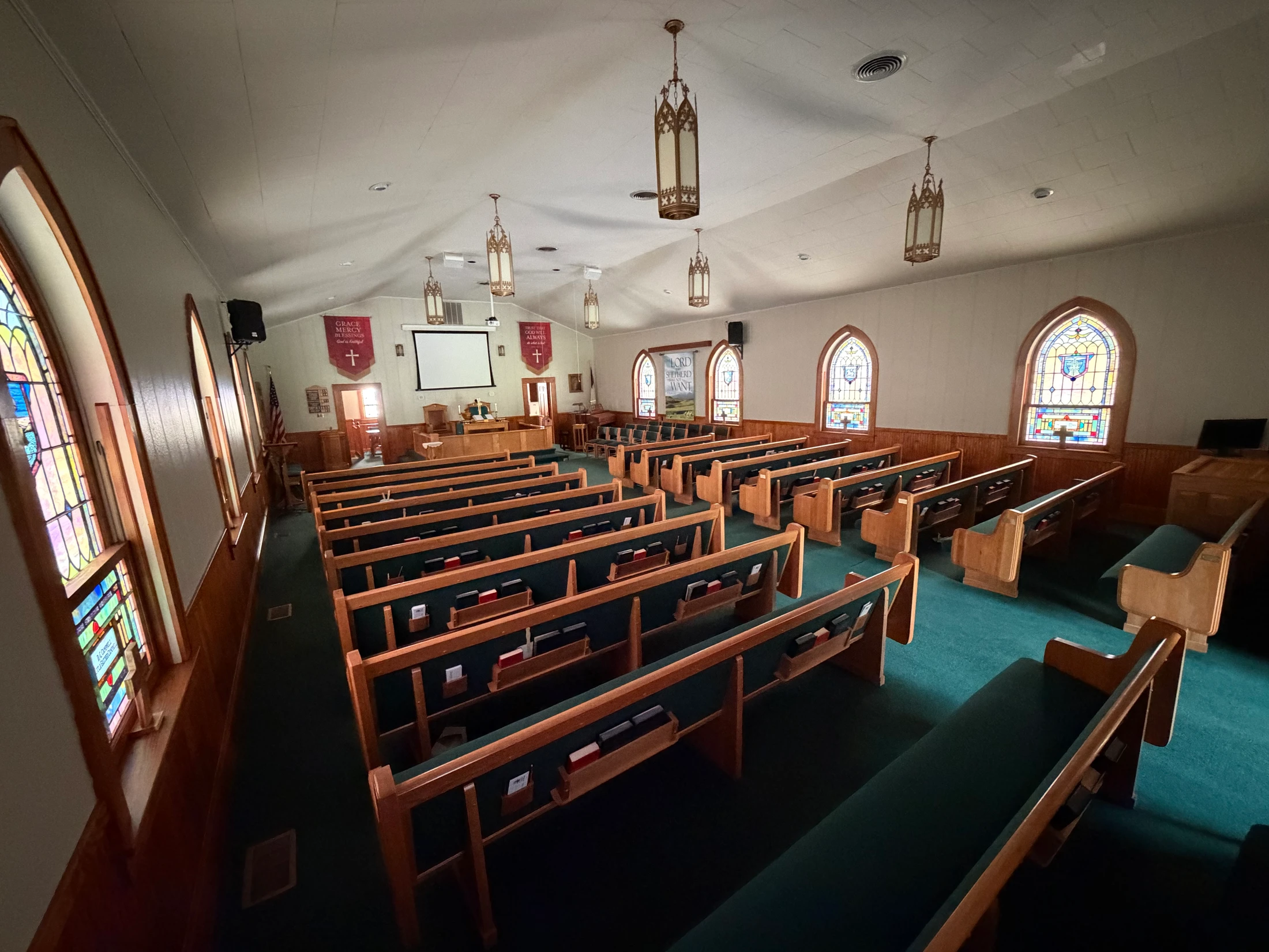A wide, high-angle view of an empty church sanctuary with wooden pews, green carpet, stained glass windows, and a pulpit at the front.