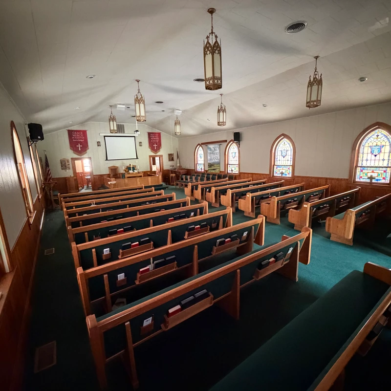 A wide, high-angle view of an empty church sanctuary with wooden pews, green carpet, stained glass windows, and a pulpit at the front.