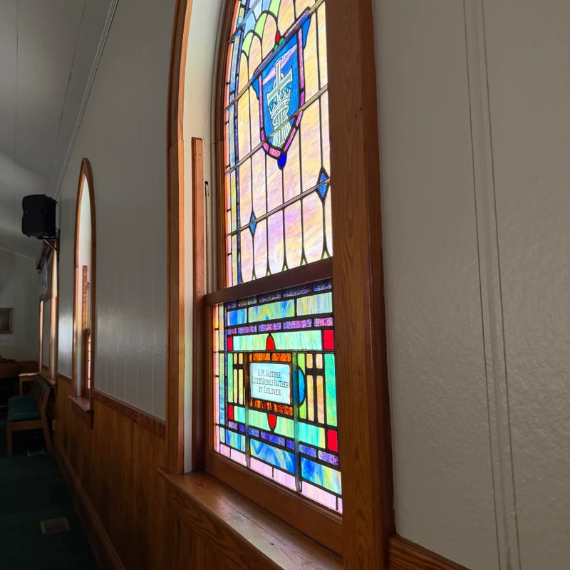 A colorful arched stained glass window in a wooden frame, letting sunlight into a church with white walls and wood paneling on the lower half.