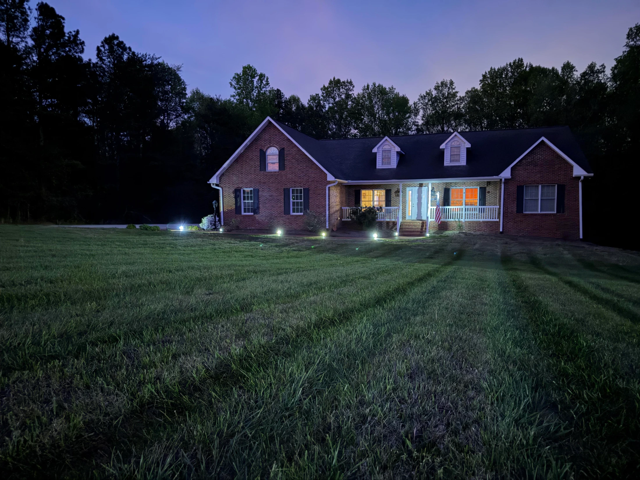 A large brick house sits on a grassy lawn at dusk. The home's windows are warmly lit, and landscape lights cast long shadows across the yard.