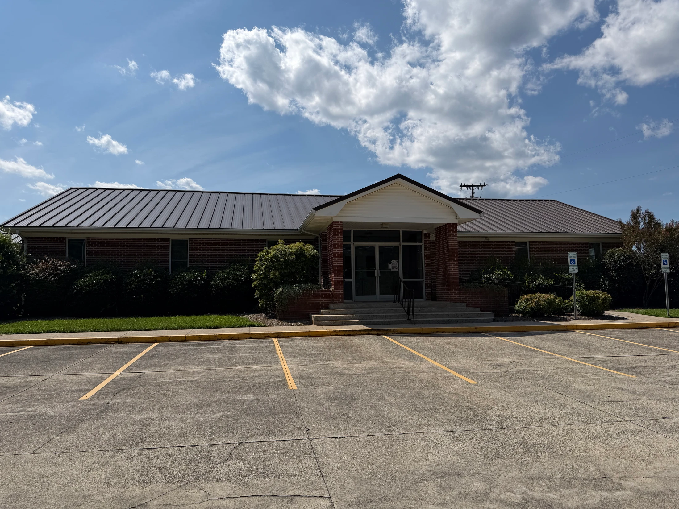 A single-story brick building with a brown metal roof, seen from an empty parking lot under a blue sky with white clouds on a sunny day.
