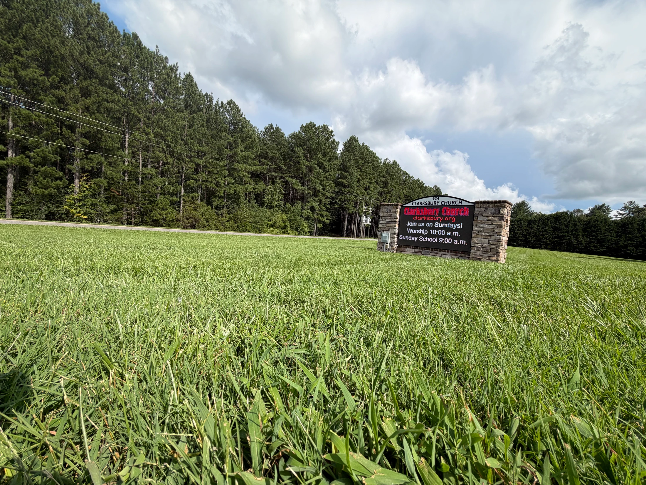 A low-angle view of a vast green lawn with a stone sign for Clarksbury Church. A line of tall pine trees stands behind the sign under a cloudy blue sky.