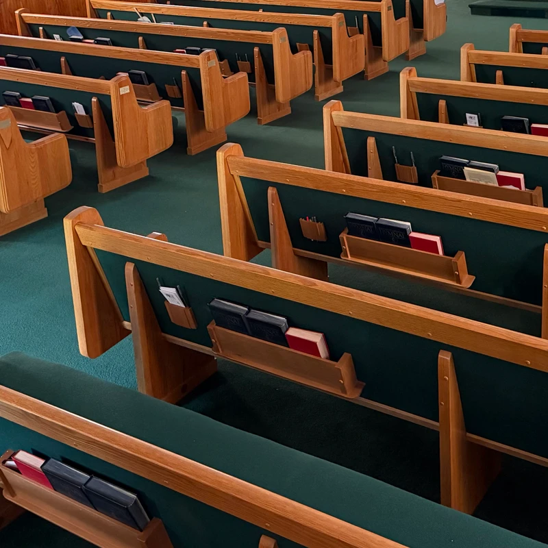 A high-angle view of an empty church shows neat rows of wooden pews with green cushions on a green carpeted floor, each pew holding hymnals.