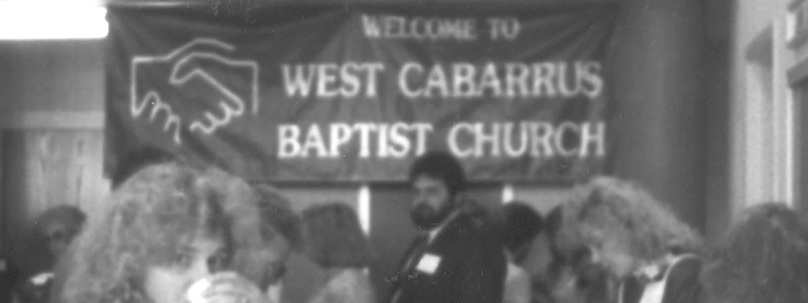 A black and white photograph of a gathering. In the background, a banner reads 'Welcome to West Cabarrus Baptist Church' with a handshake logo.