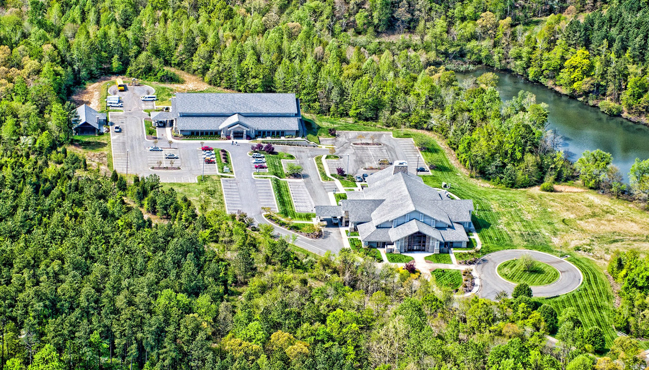 An aerial shot of two large, modern church buildings with gray roofs, set in a clearing in a dense green forest next to a winding river.
