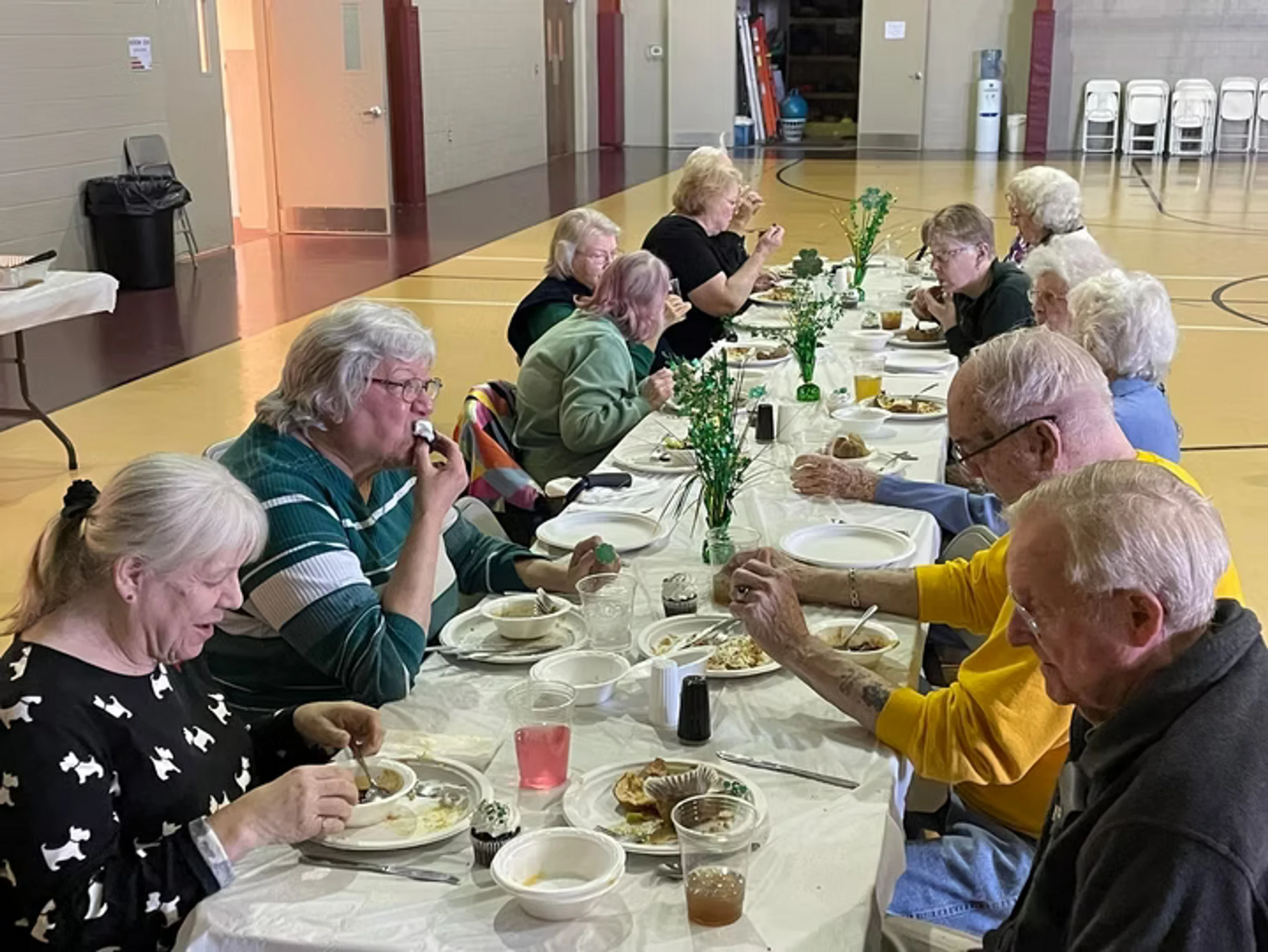A group of seniors are seated at a long table covered with a white cloth in a gymnasium, eating a meal together with green St. Patrick's Day decorations.