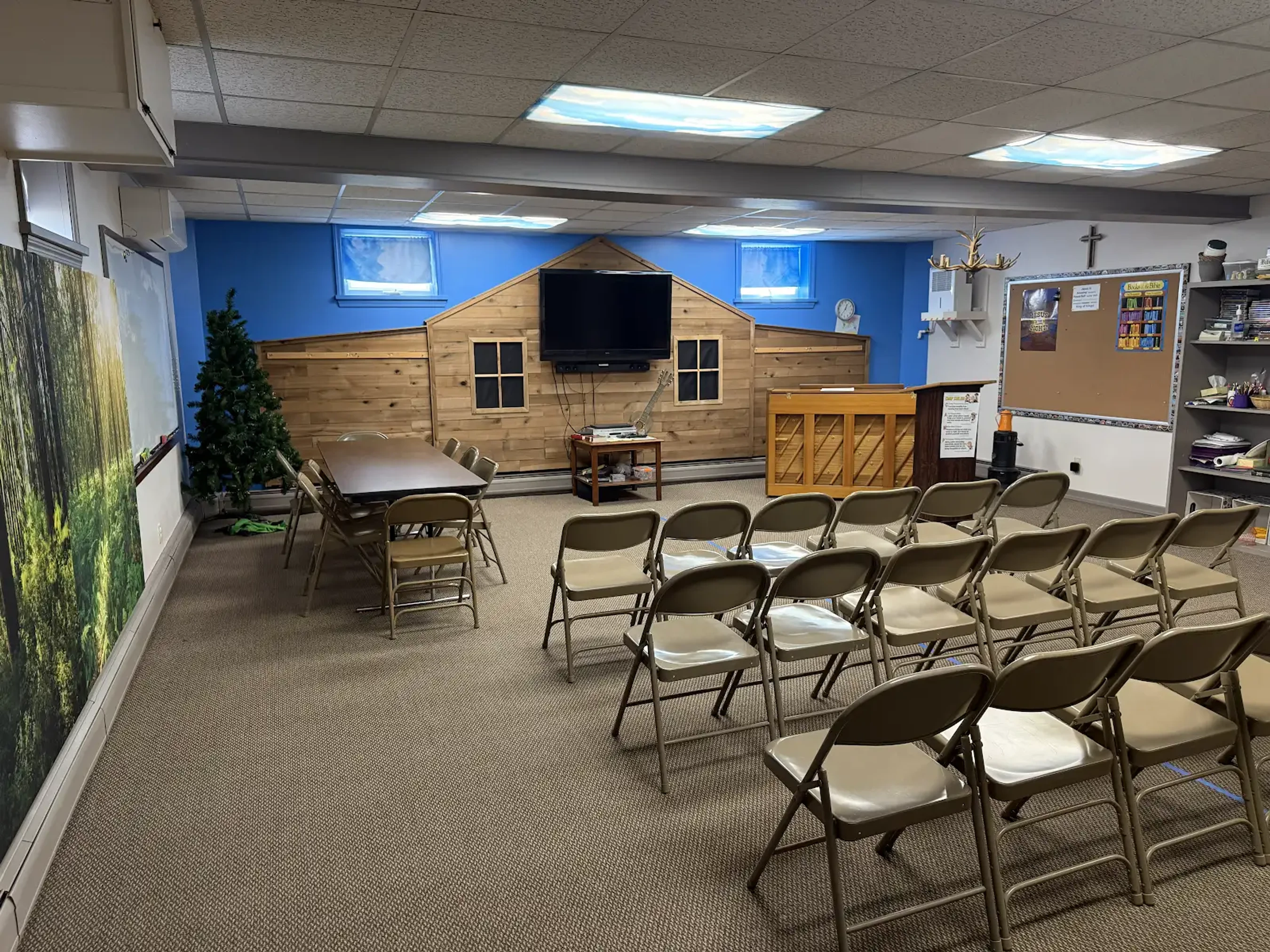 A church meeting room with rows of metal folding chairs facing a blue wall with a wooden house-like facade, a TV, a piano, and a podium.