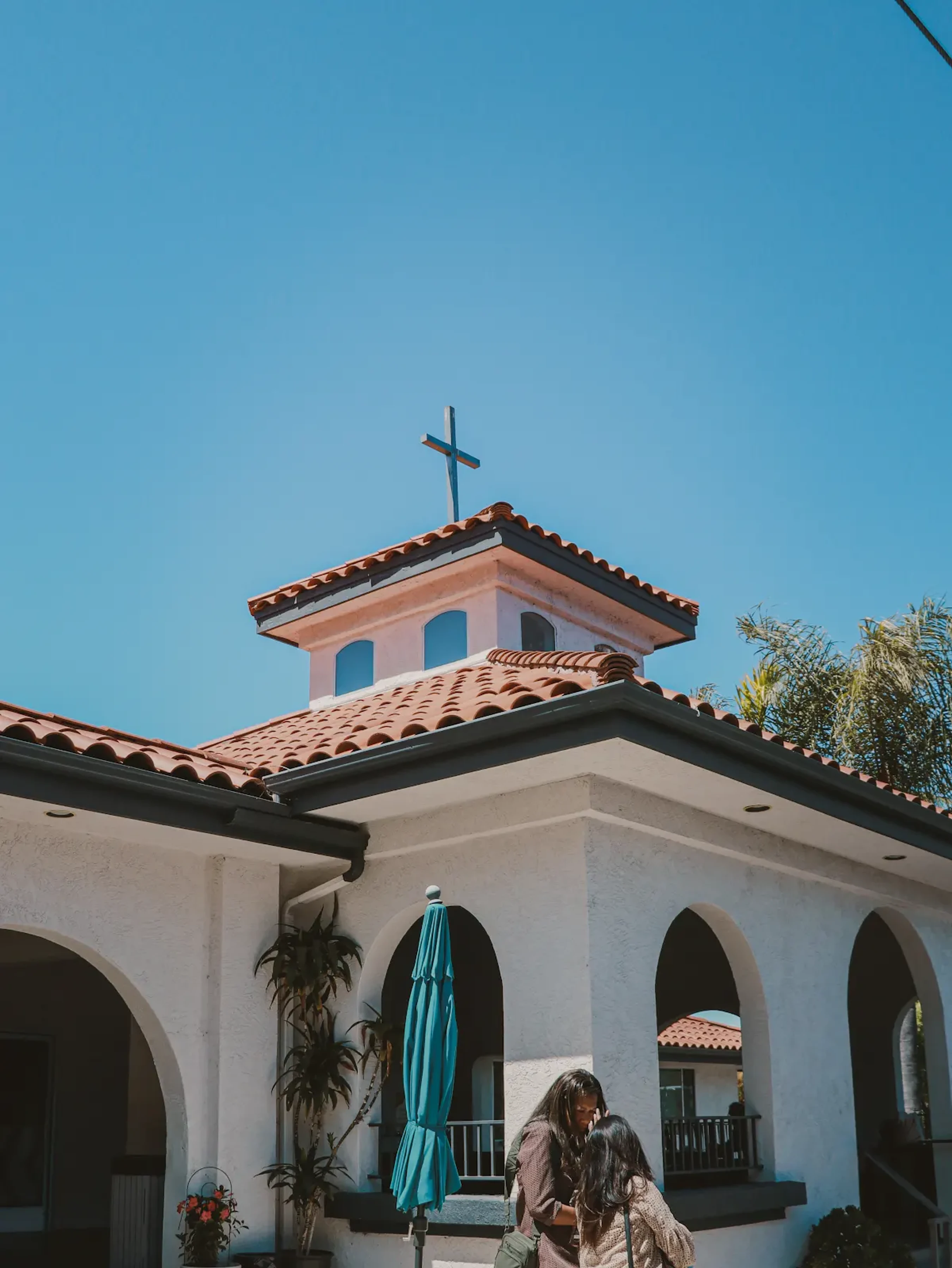 A white, Spanish-style church with a red tile roof and a cross on top, under a clear blue sky. Two people stand together in the foreground.