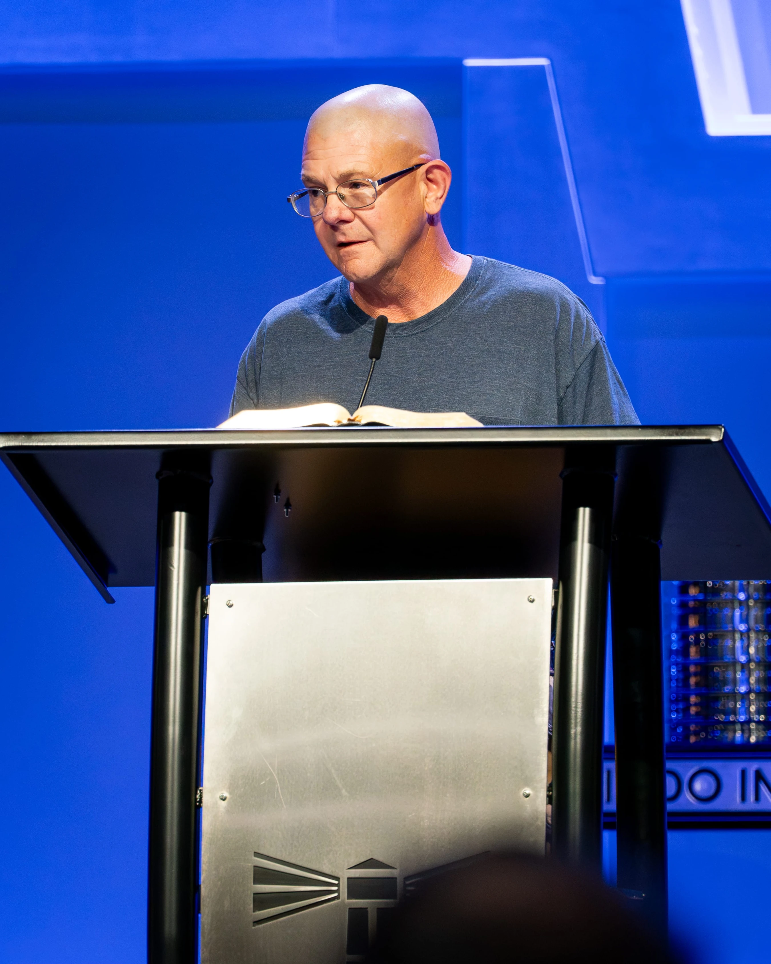 A bald man with glasses wearing a grey t-shirt speaks at a lectern. An open book and a microphone are in front of him against a bright blue background.