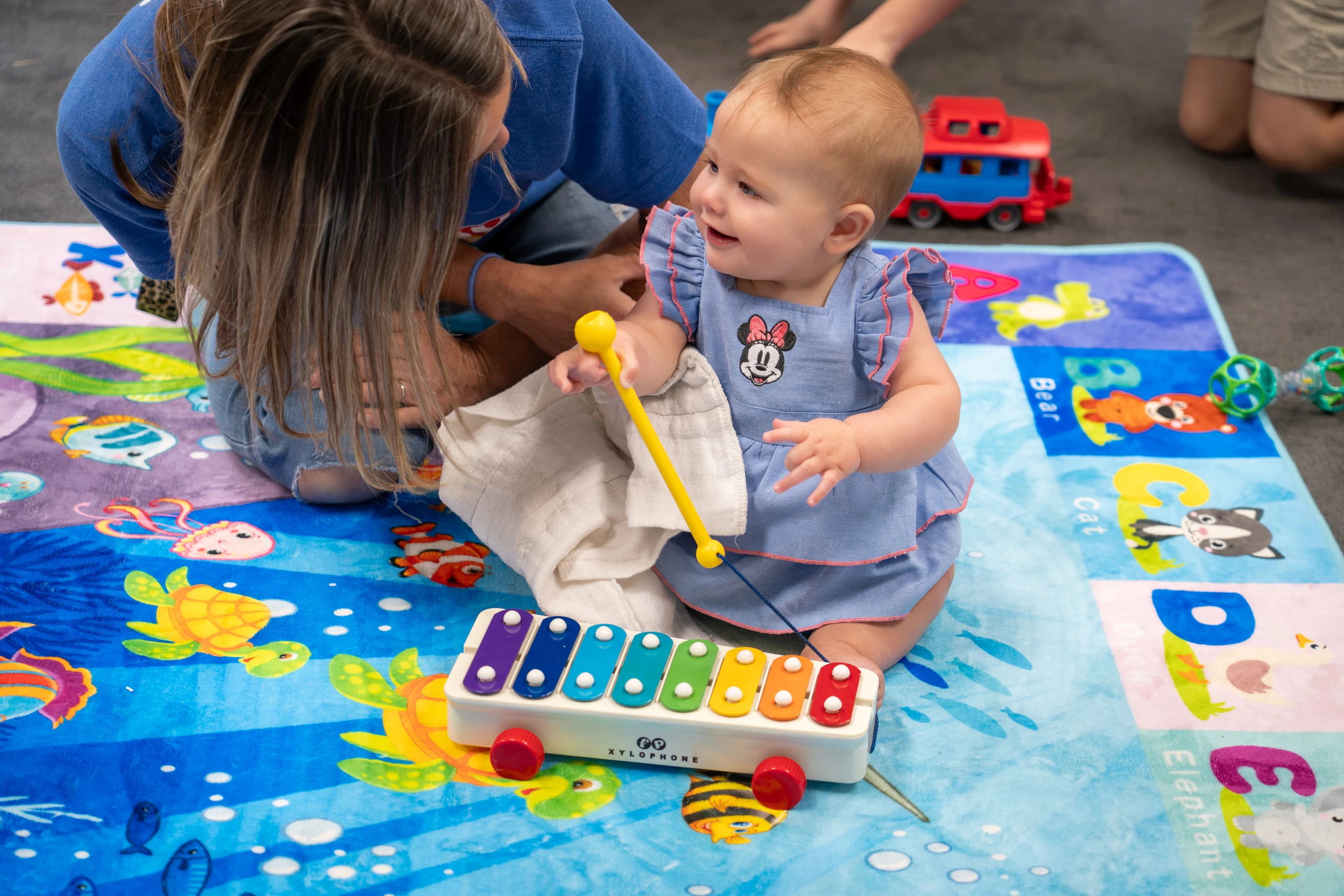 A smiling baby girl in a blue dress sits on a colorful playmat, holding a yellow mallet over a toy xylophone as a woman leans in towards her.