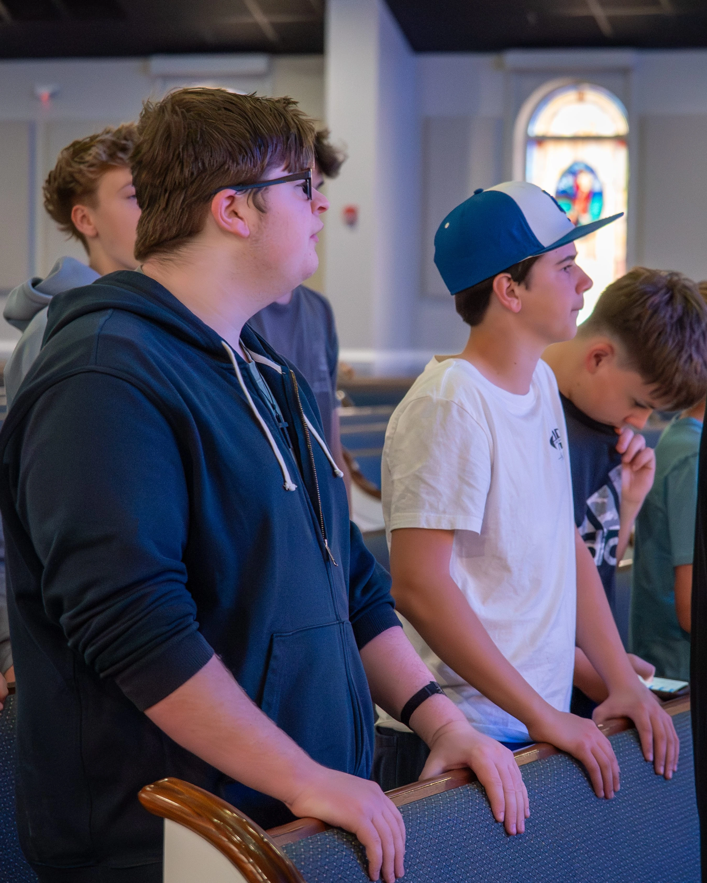 A group of teenage boys stand in a row in a church sanctuary, resting their hands on the pew in front of them while looking forward attentively.