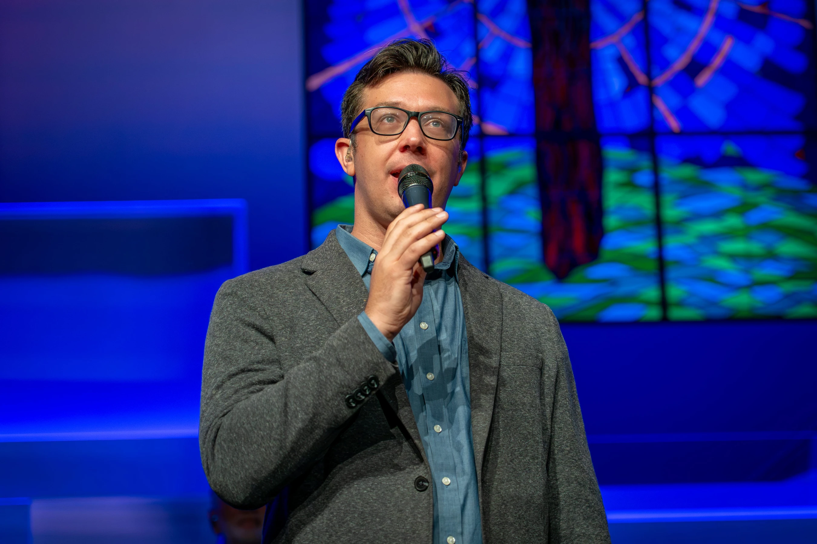 A man wearing glasses and a grey blazer speaks into a microphone on a stage. The background is lit with blue light and features a colorful abstract design.