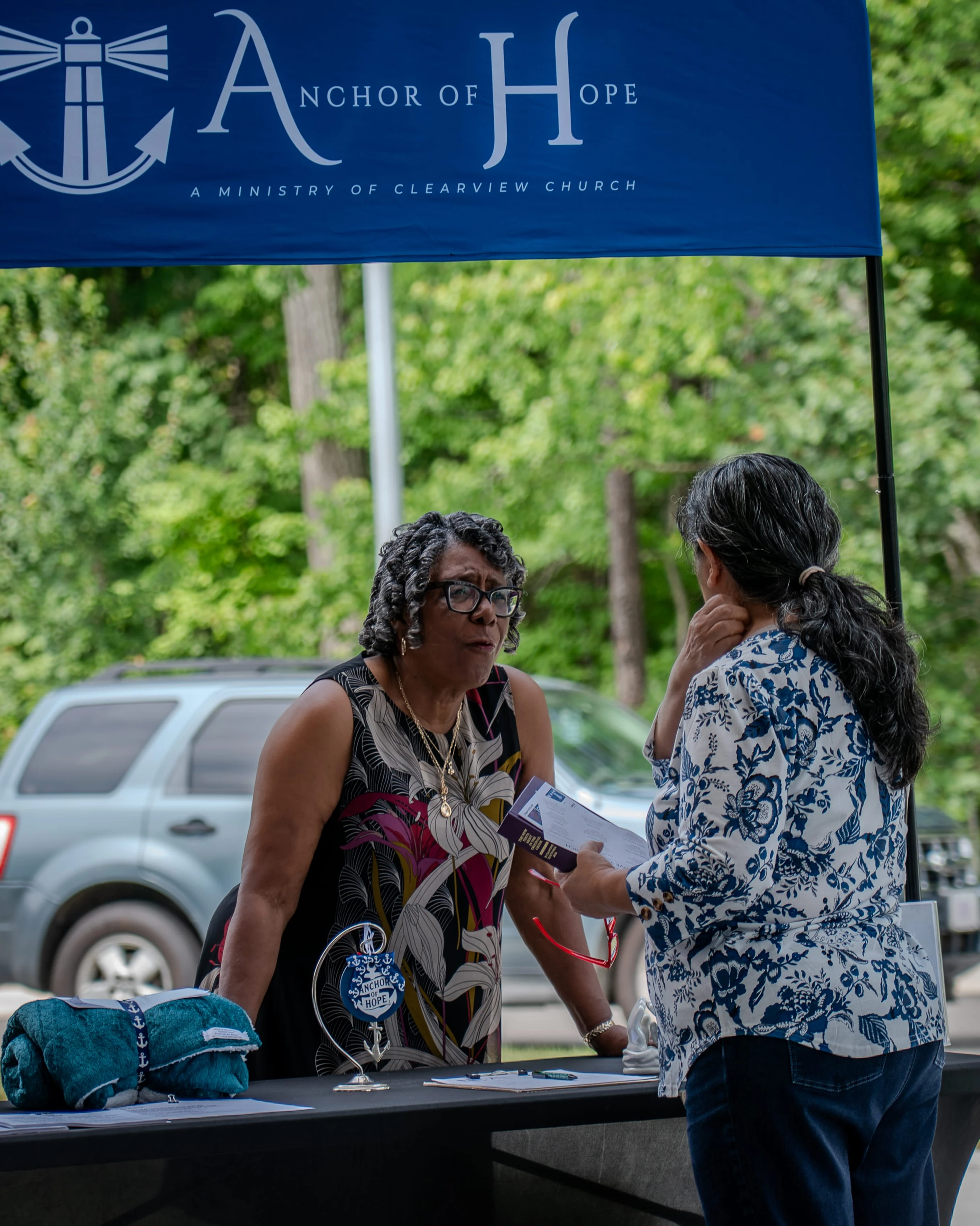 Two women talk at a table under a blue banner for the 'Anchor of Hope' ministry at an outdoor event. One woman listens while the other speaks.