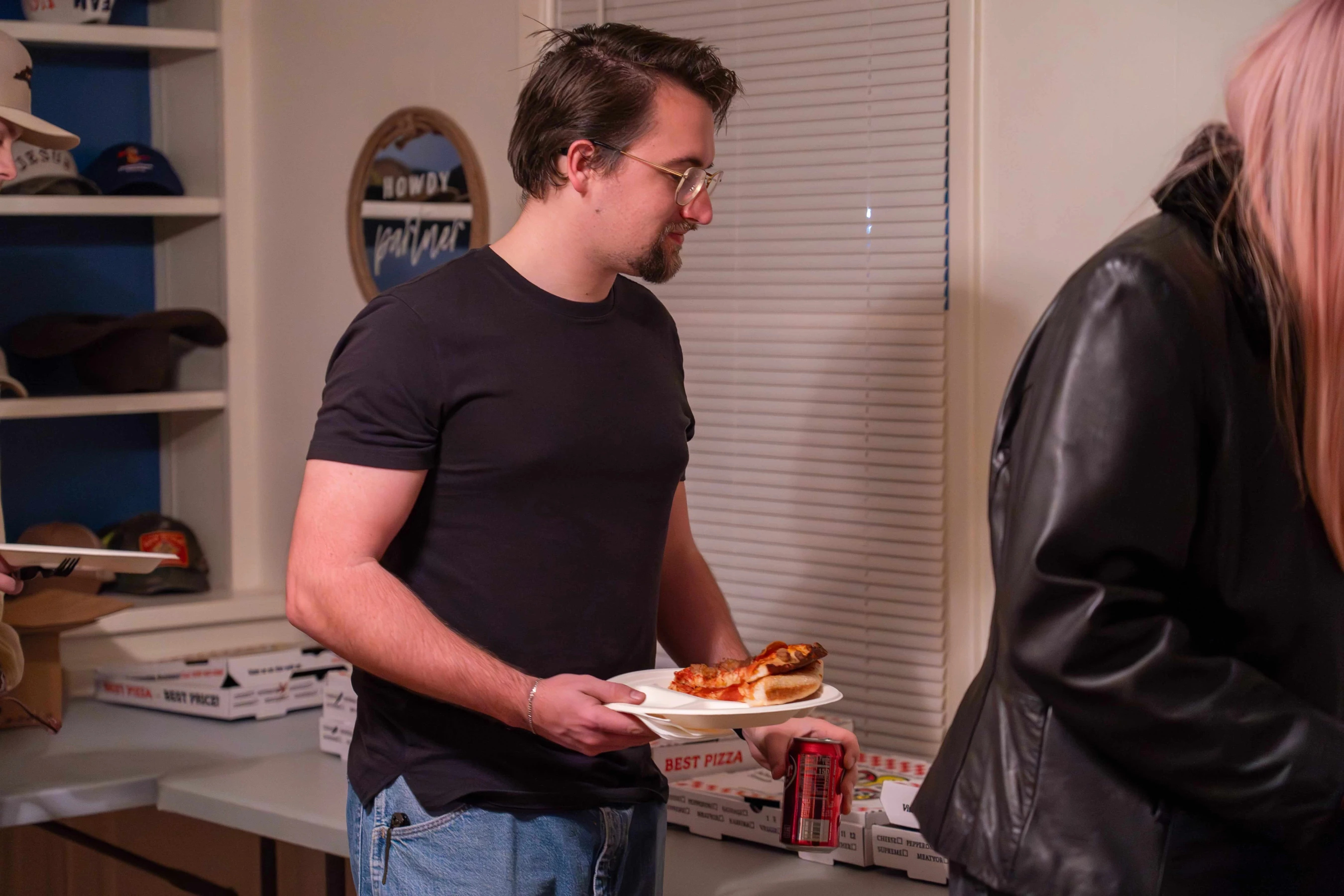 A man with glasses and a black t-shirt holds a paper plate with pizza slices. He stands in front of a table with stacks of pizza boxes.