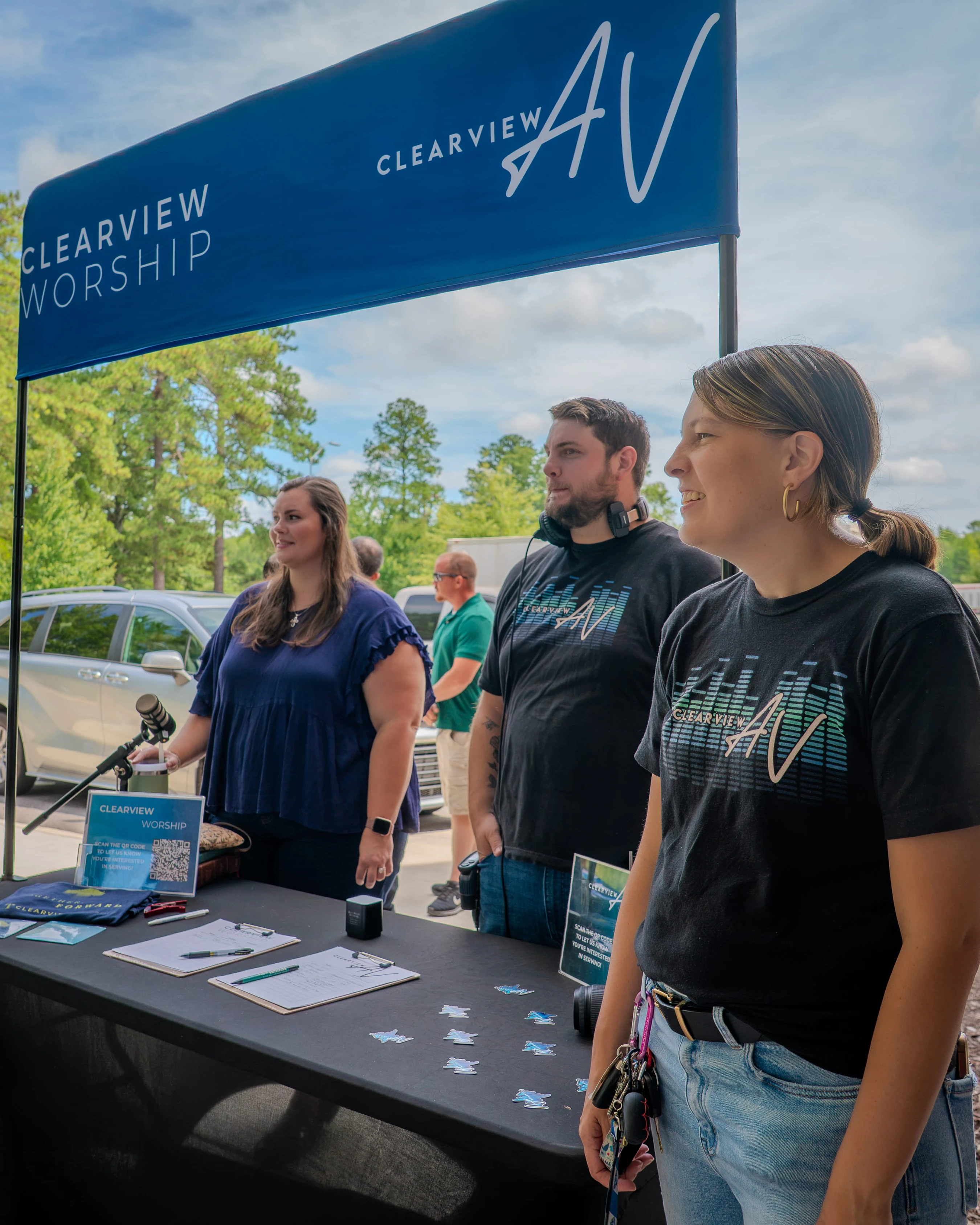 A man and two women from the Clearview AV and Worship teams stand at an outdoor information booth, ready to greet visitors on a sunny day.