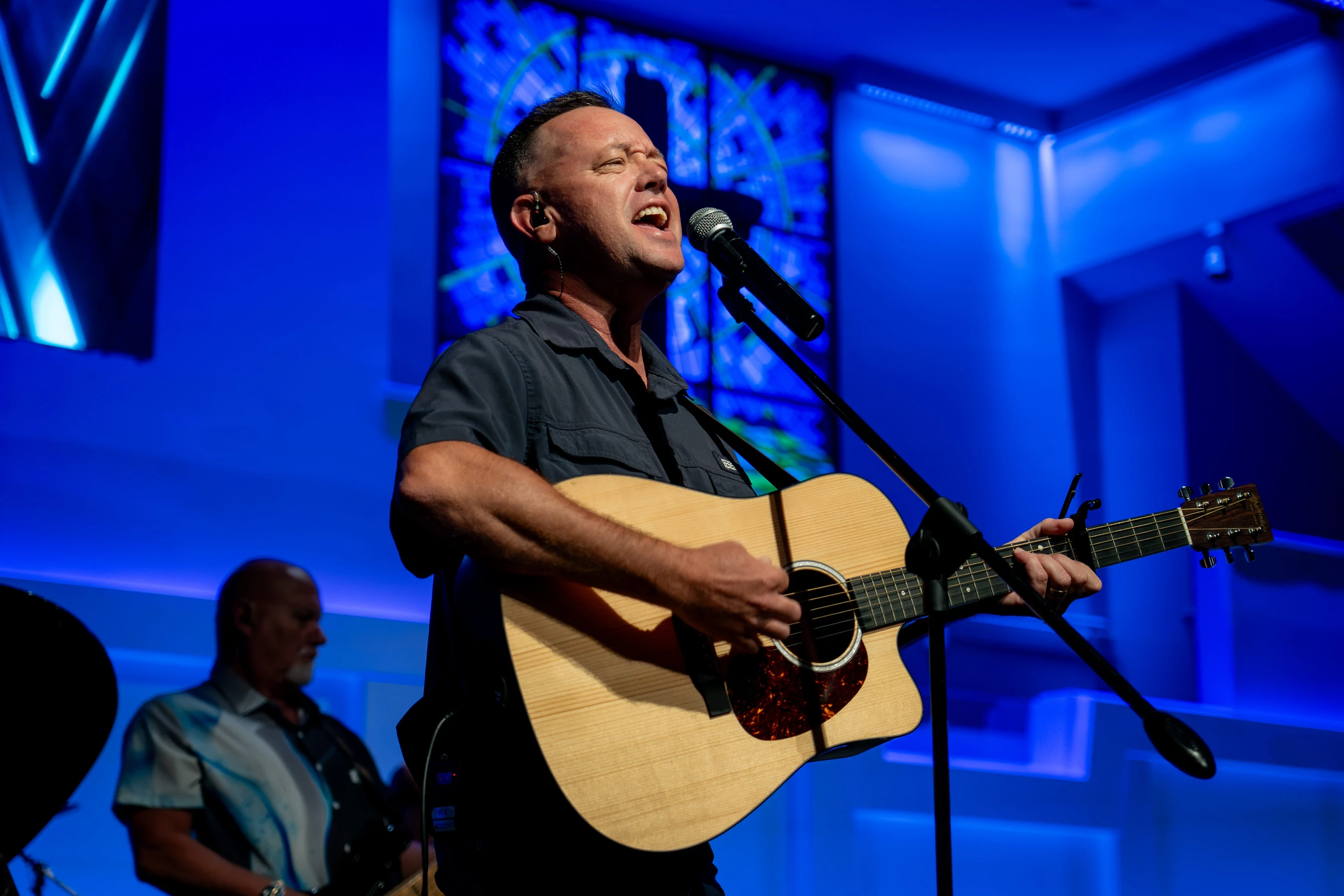 A man sings passionately into a microphone while playing an acoustic guitar on a stage bathed in vibrant blue light, with another musician in the background.