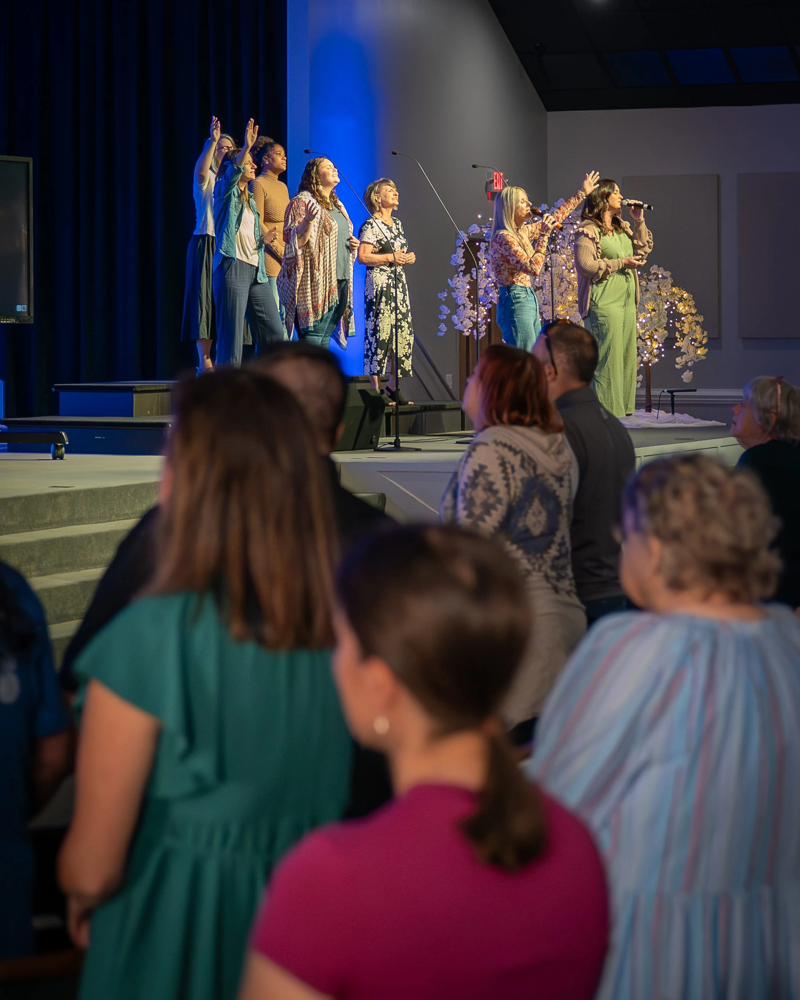 A group of women sing on a stage with blue backlighting, leading a congregation in worship. The audience is seen from behind, looking toward the stage.