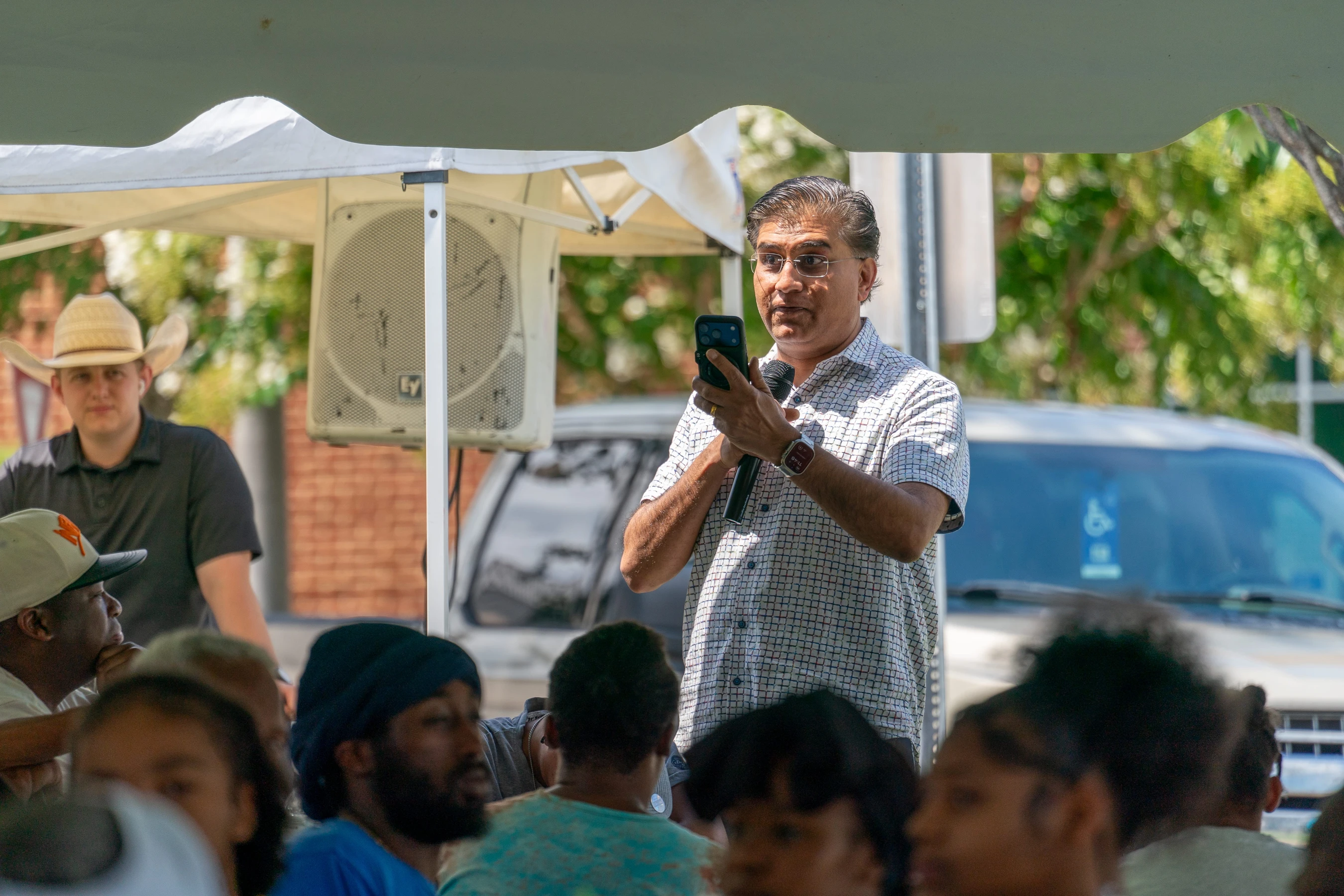 A man with glasses stands under a canopy tent, speaking into a microphone and looking at his phone while addressing a seated crowd at an outdoor event.