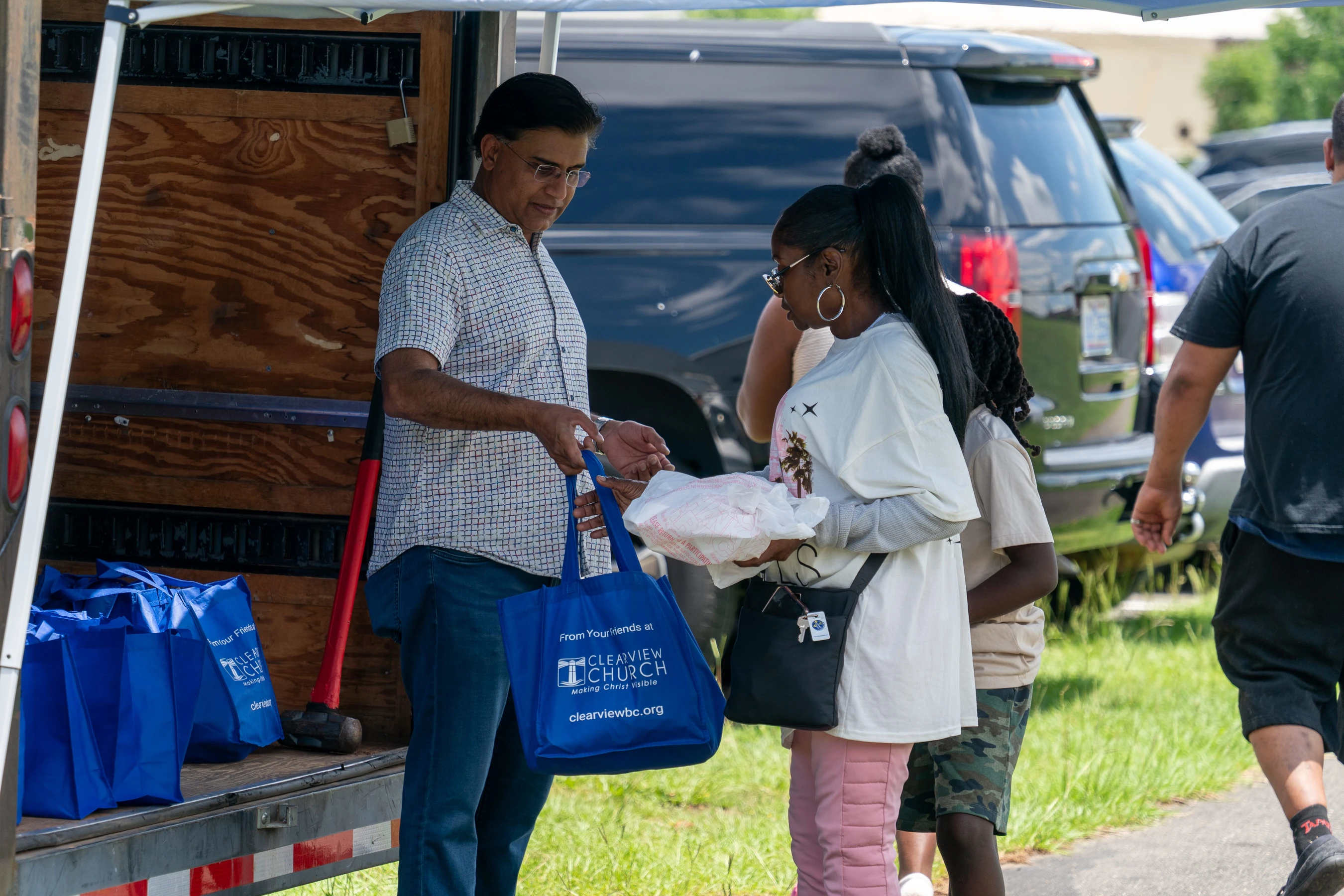 A man in a patterned shirt stands by a truck and hands a blue tote bag with a church logo to a young woman during an outdoor giveaway event.