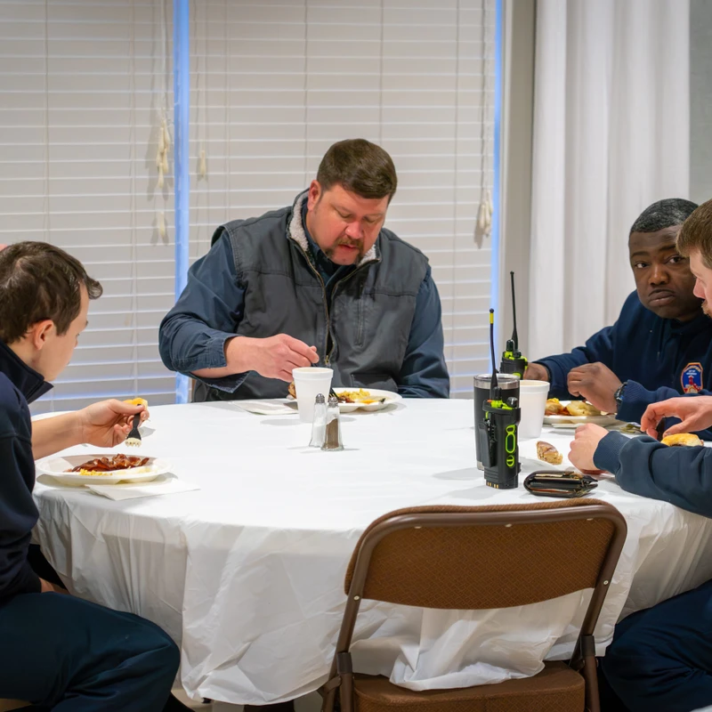 Five firefighters in dark blue uniforms sit around a round table with a white cloth, eating a meal together from paper plates in a brightly lit room.