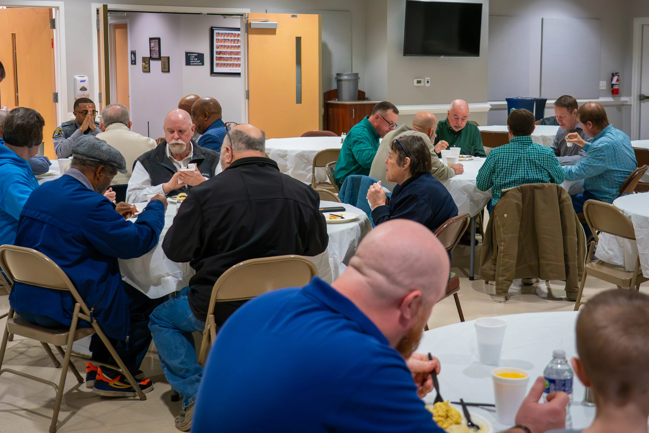 A diverse group of men and a woman sit at round tables with white cloths, eating breakfast and talking together in a community hall.