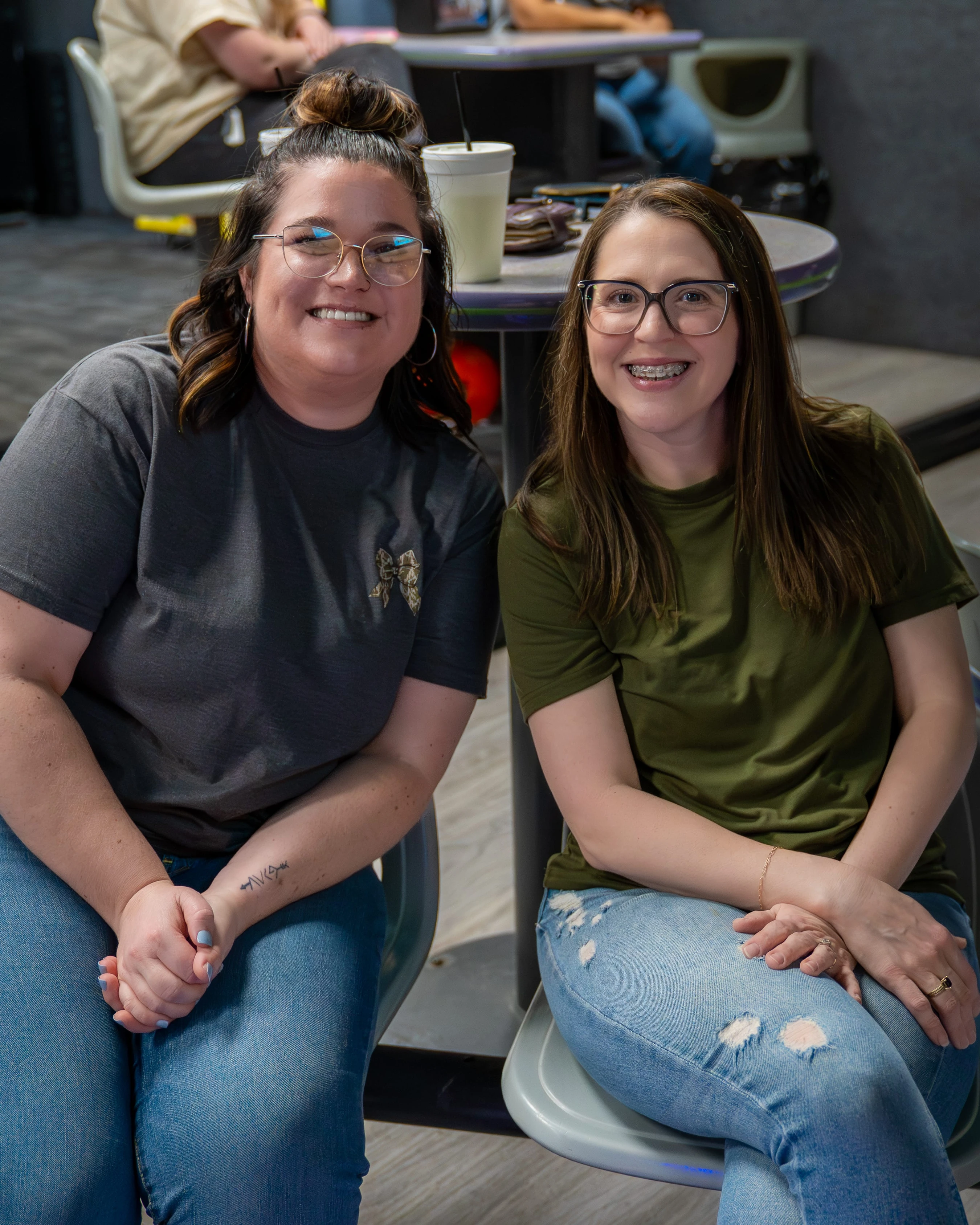 Two women wearing glasses and casual clothing sit side-by-side in chairs at what appears to be a bowling alley, both smiling warmly at the camera.