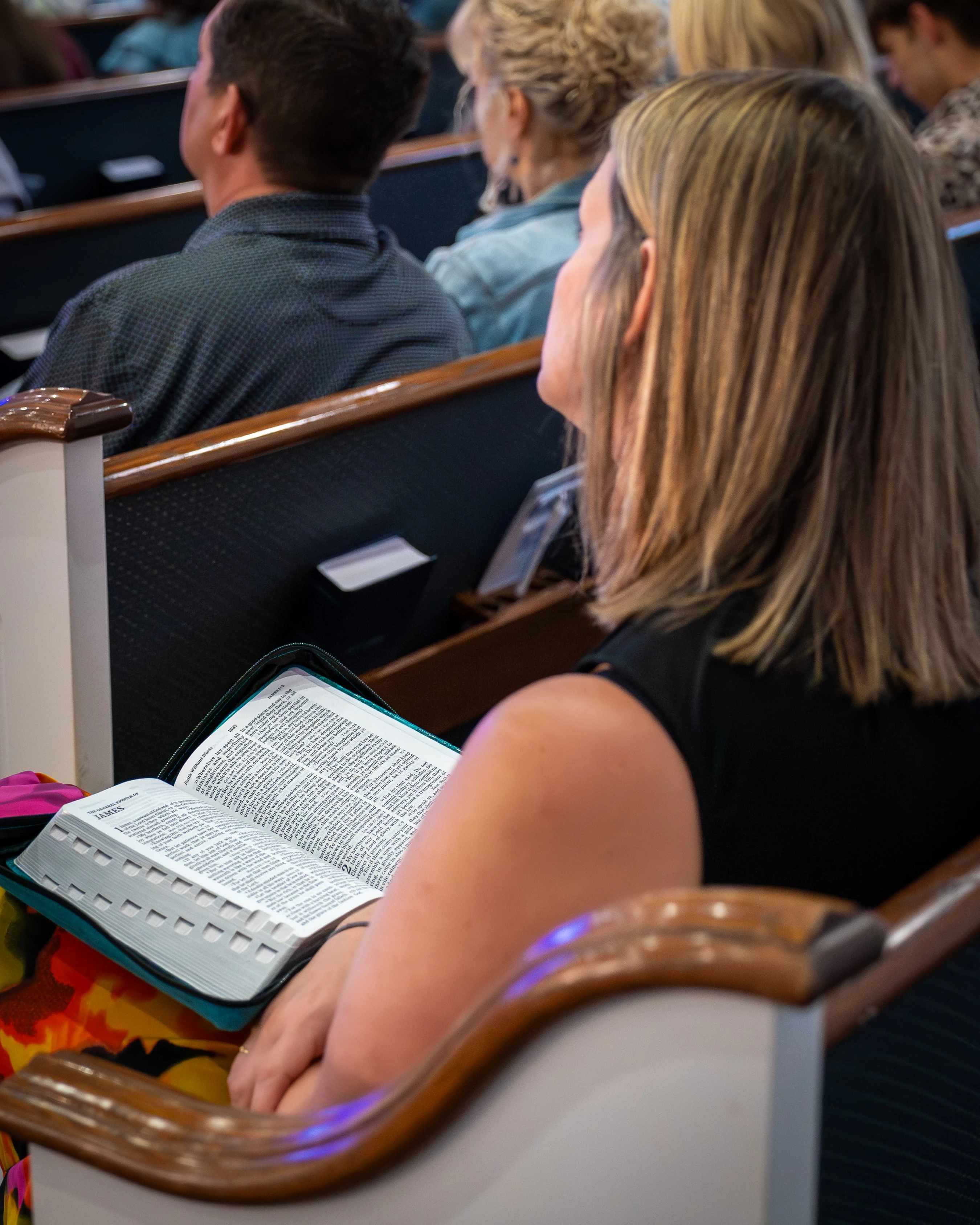 A blonde woman sits in a church pew, holding an open Bible in her lap. She is looking forward, listening to a service with other congregants.