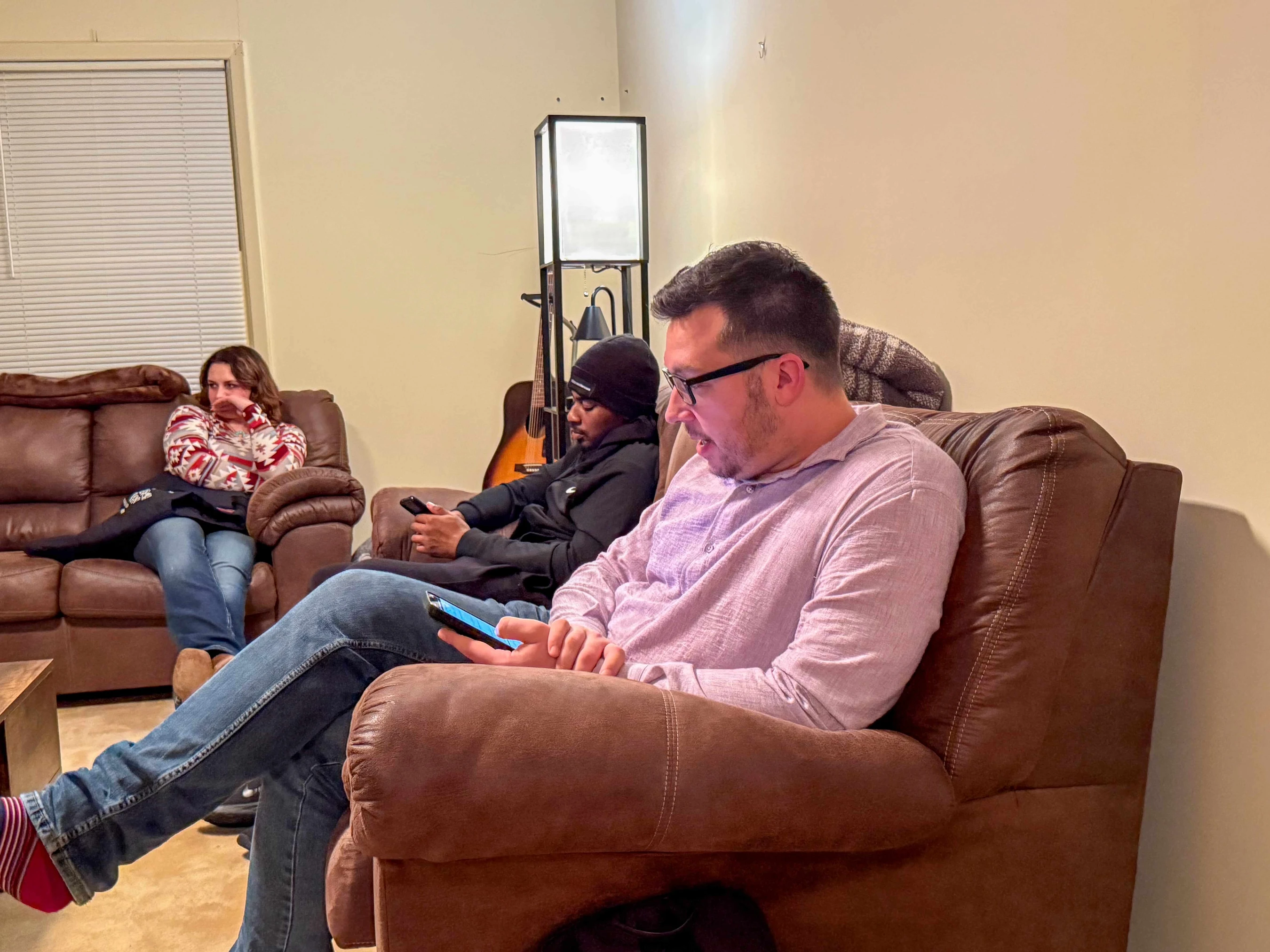 Three friends relax on brown couches in a living room. Two men are focused on their phones, while a woman in the background looks off thoughtfully.