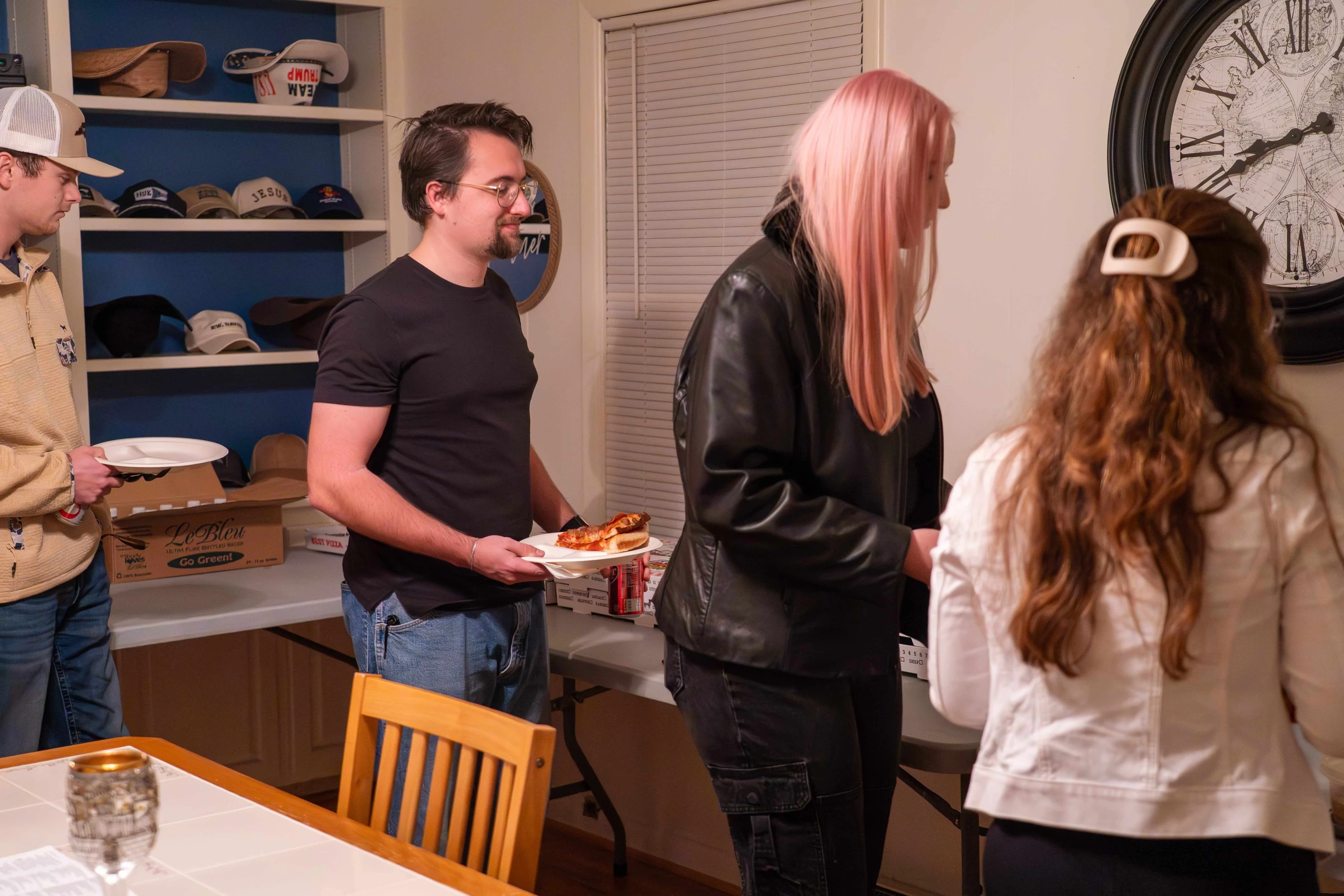 A group of young people stand in a line at a casual house party, serving themselves pizza from boxes on a long table. A man in a black shirt smiles.