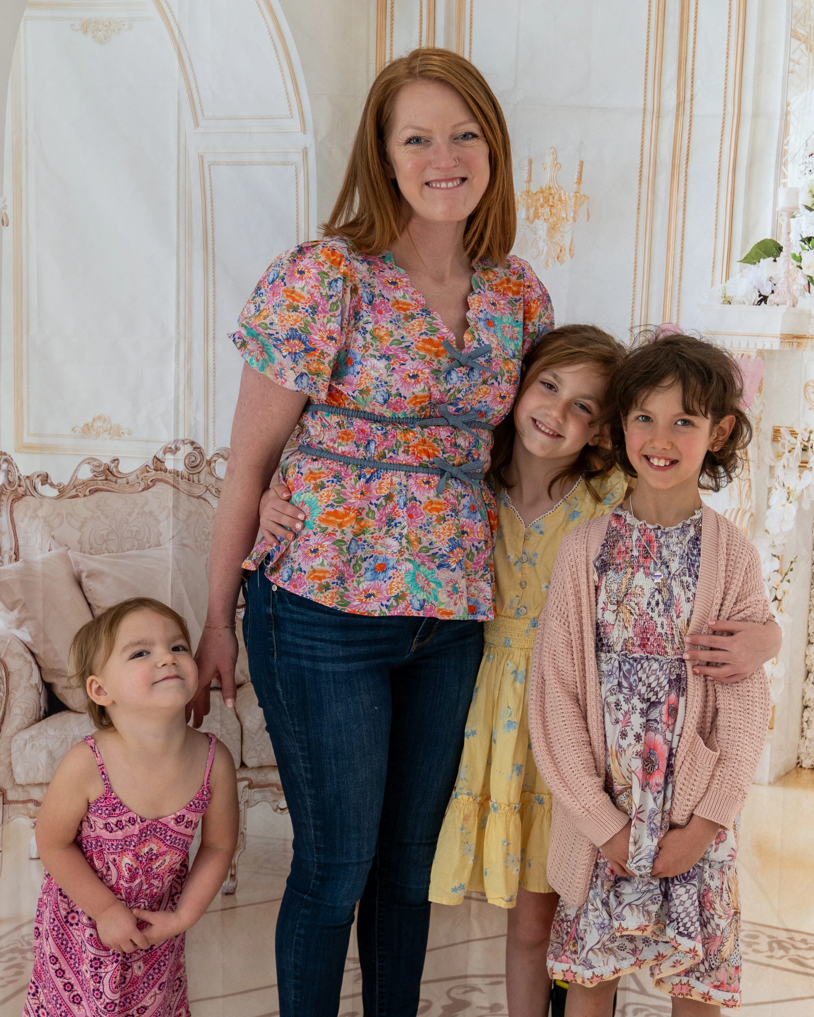 A smiling red-haired woman poses with her three young daughters against an ornate backdrop. They are all looking at the camera and smiling.