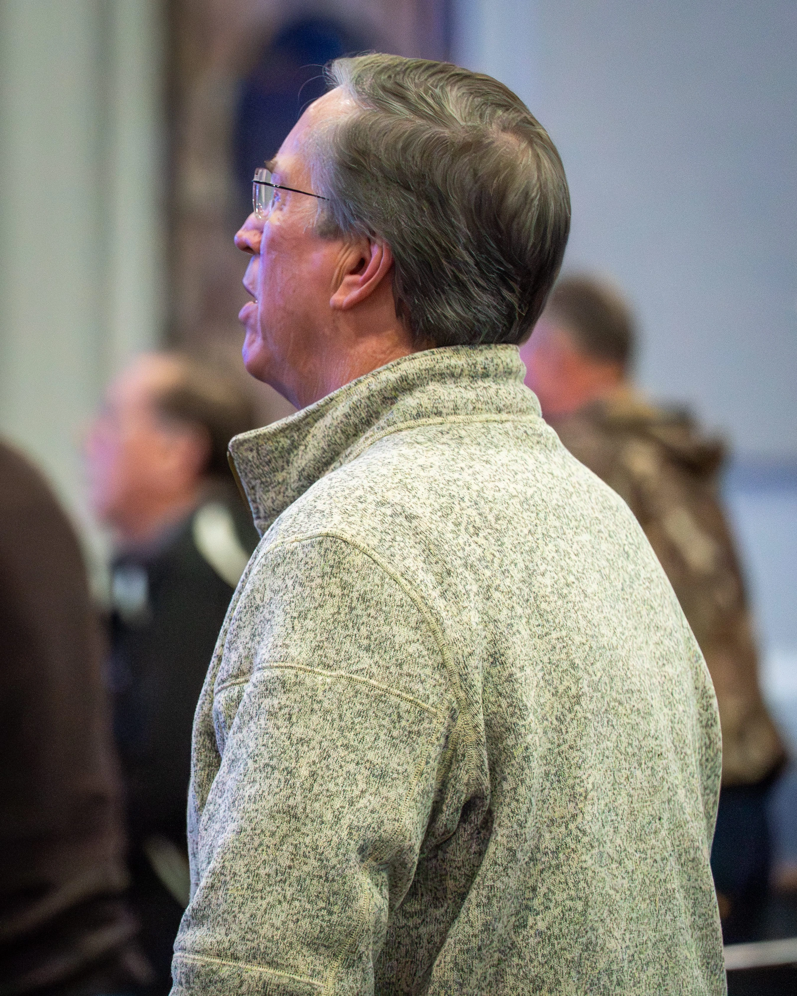 A side-profile view from behind of an older man with glasses and grey hair, wearing a flecked grey fleece pullover. He is looking up and to the left.