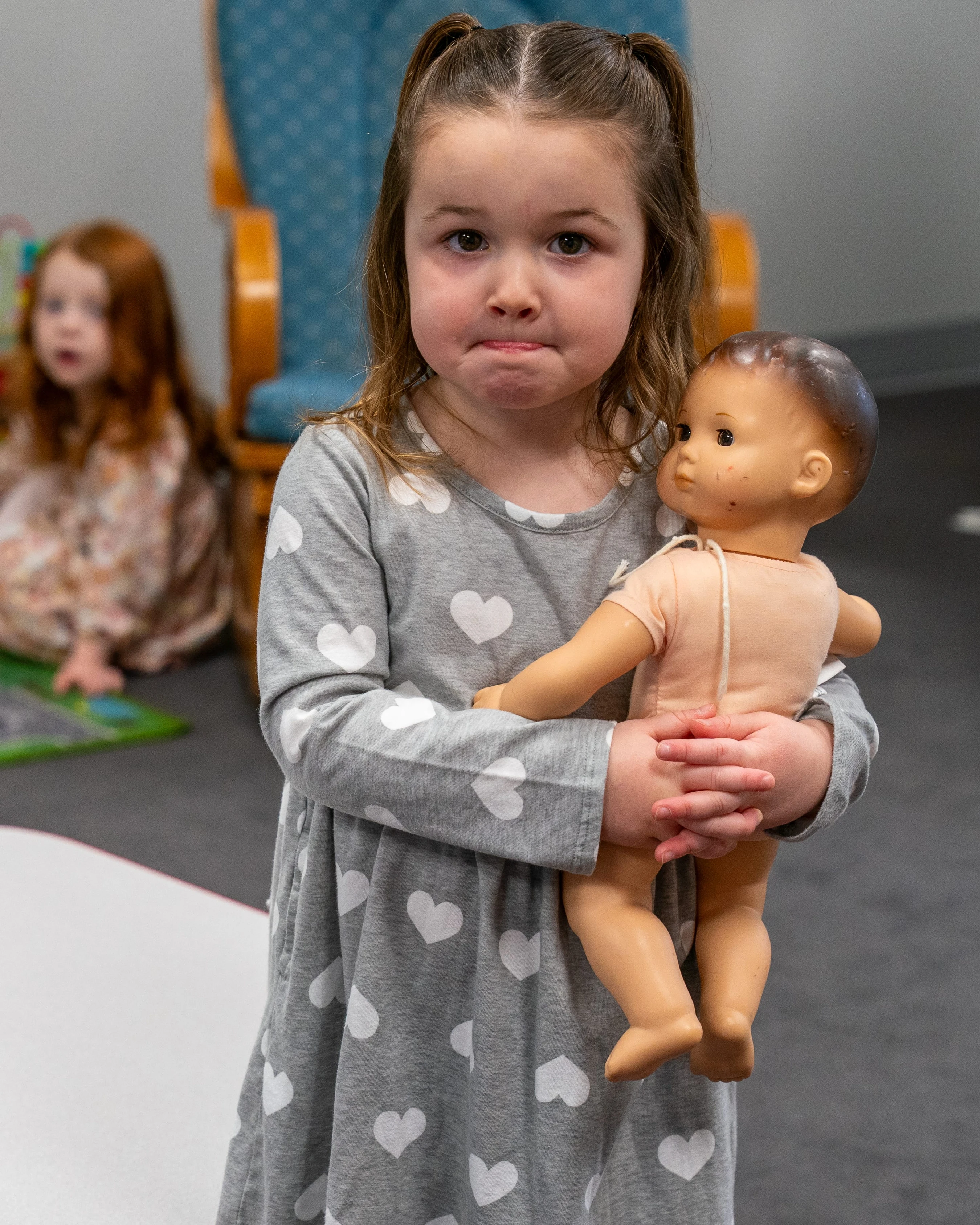 A young girl with a pouting face, wearing a grey dress with white hearts, stands holding a vintage baby doll. Another child sits in the background.