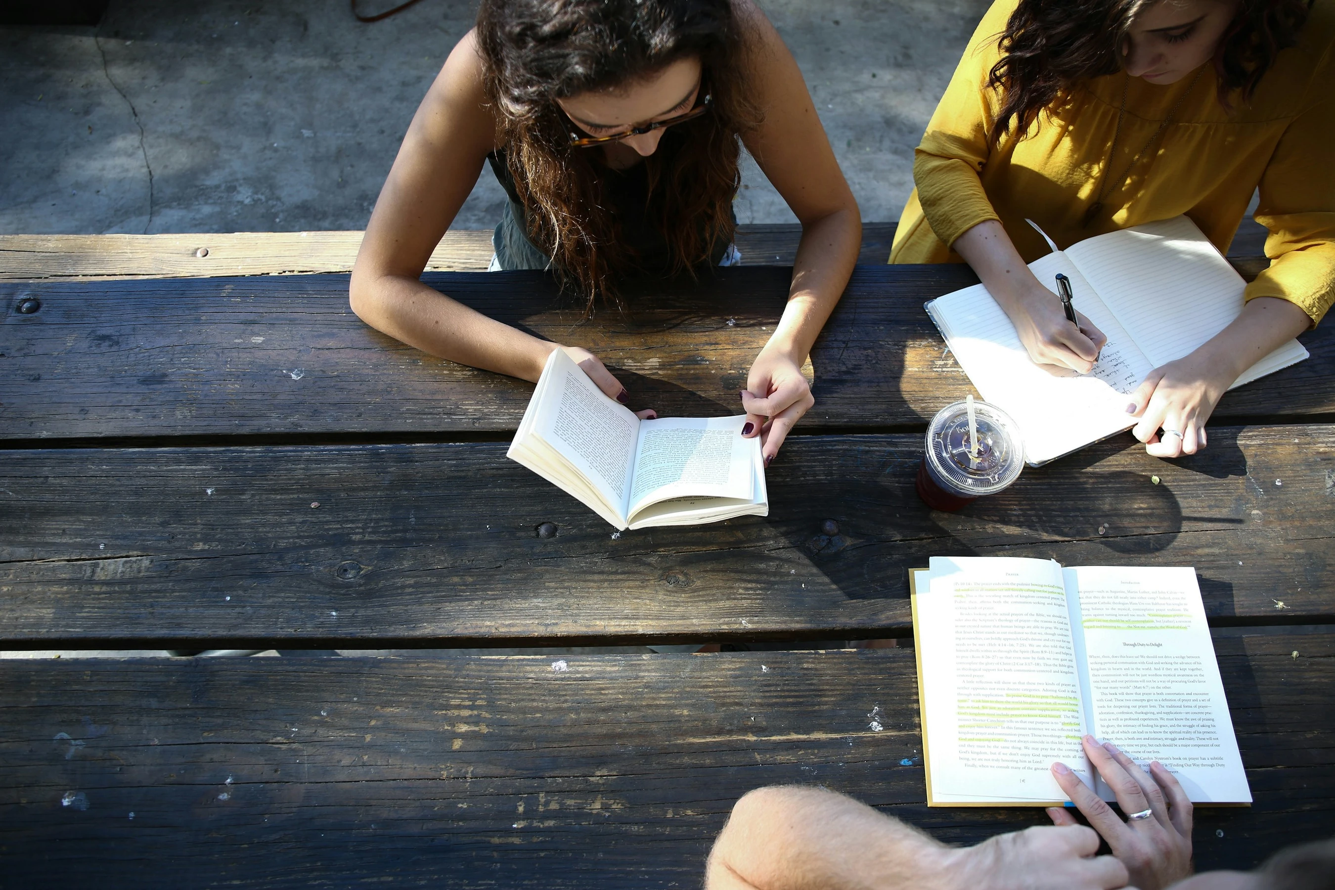 Two people reading books and writing in a notebook at a wooden picnic table