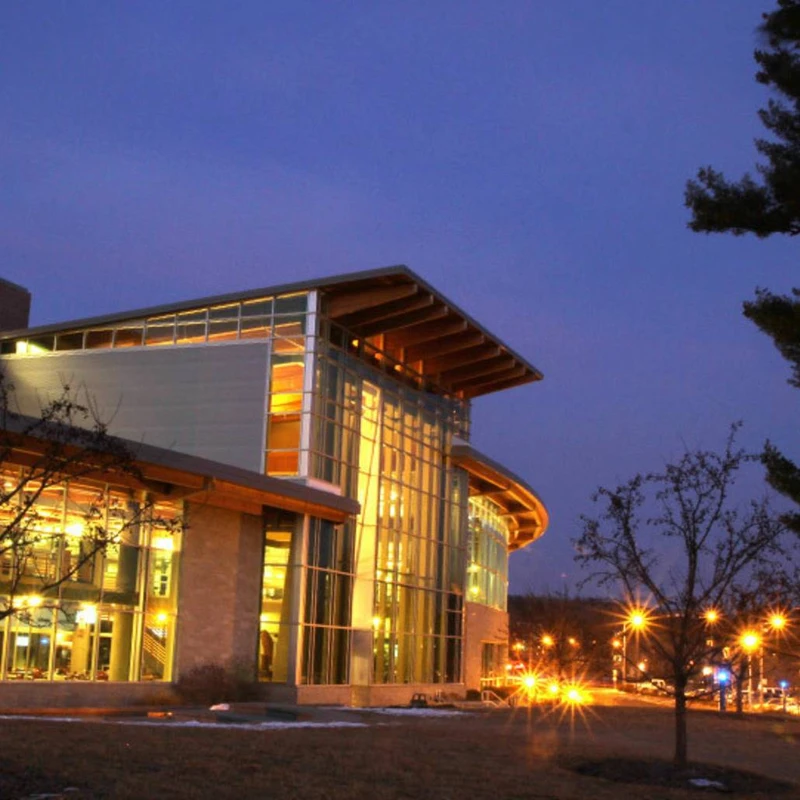 A modern building with large, illuminated glass windows glows warmly against a deep blue evening sky, with bare trees and streetlights in the foreground.