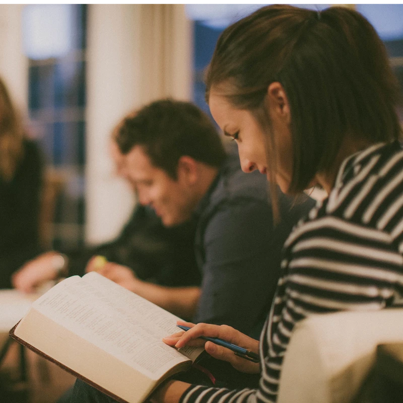 A young woman in a striped shirt reads a large book while holding a pen. In the background, other people are also studying in a softly lit room.