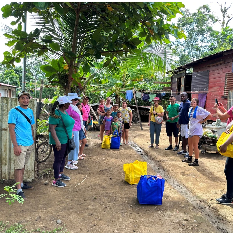 A diverse group of volunteers and local residents, including children, stand on a dirt path in a village, with yellow and blue bags of supplies between them.