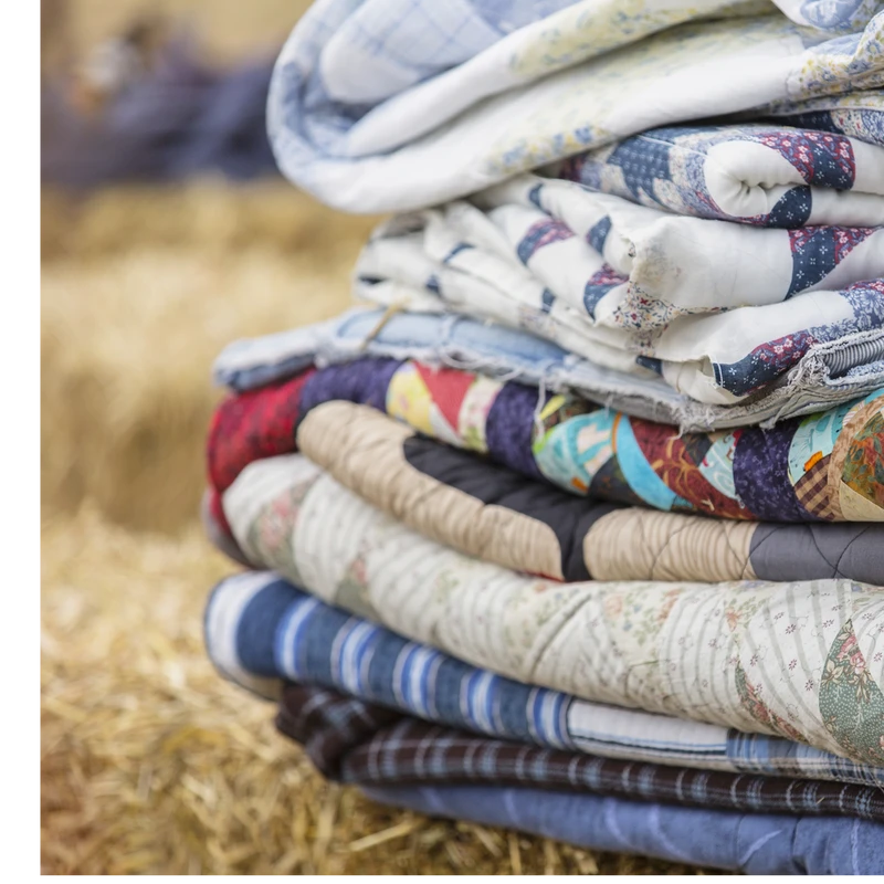 A close-up of a tall stack of folded quilts with various colors and patterns, sitting on a bed of hay with a soft-focus background.